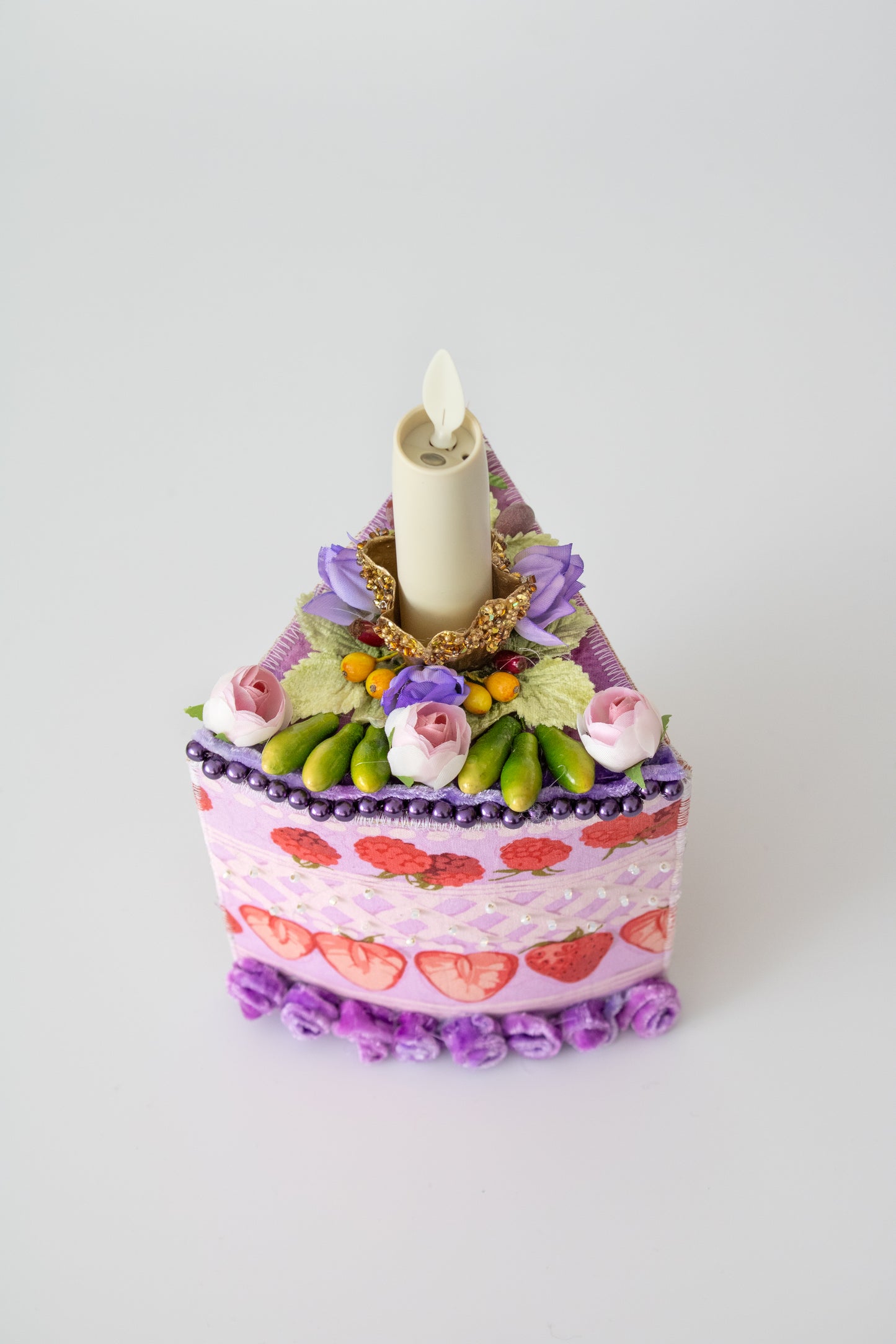 The Piece of Cake Candleholder: A DIY Tabletopper by Creative Spark is a whimsical wedge-shaped decor piece with purple and pink faux flowers, strawberries, beads, and an artificial candle on a plain white background.
