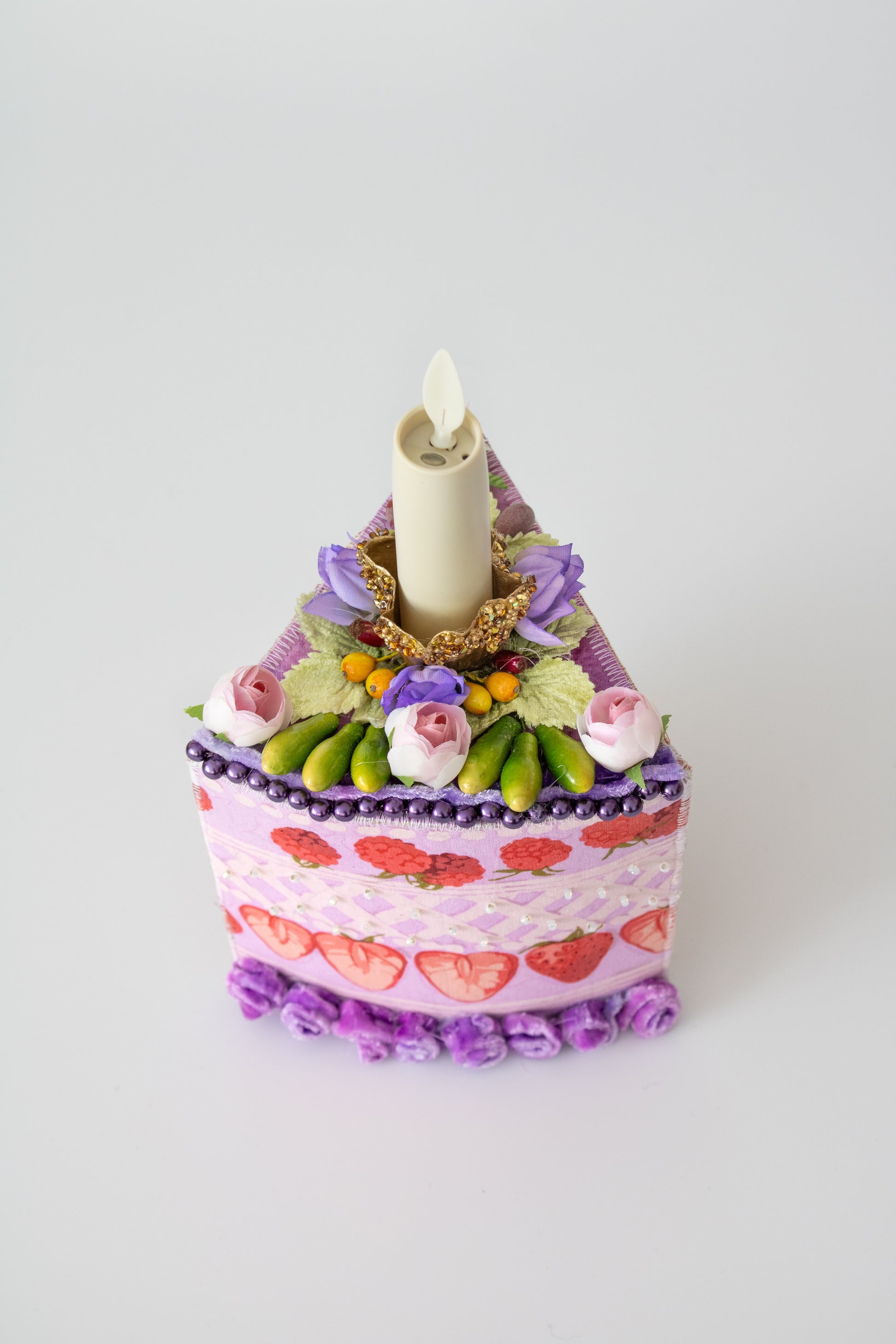 The Piece of Cake Candleholder: A DIY Tabletopper by Creative Spark is a whimsical wedge-shaped decor piece with purple and pink faux flowers, strawberries, beads, and an artificial candle on a plain white background.