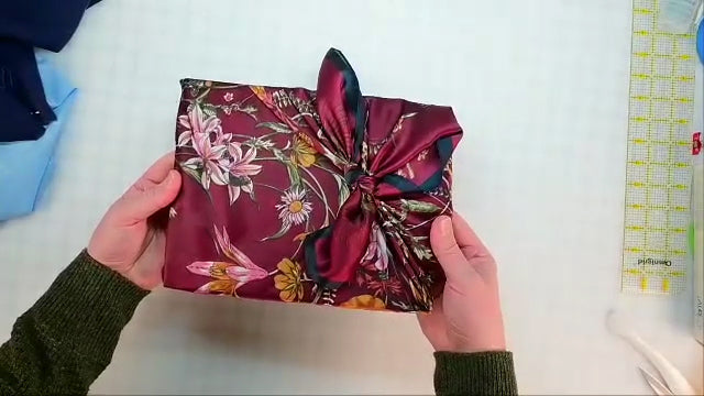 A person holds a rectangular gift wrapped in Creative Spark’s Furoshiki Wrapping: Gifts and Beyond on a white grid-patterned surface, with a yellow measuring tape and fabric scraps nearby.