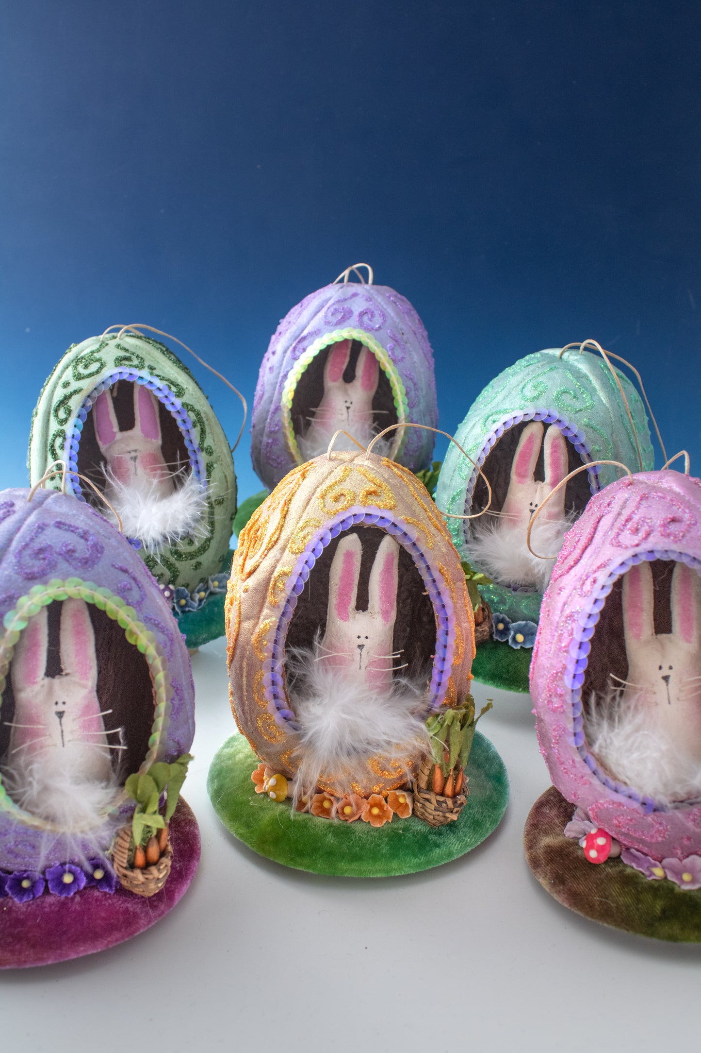 The Creative Spark Easter Egg-stravaganza: DIY Light-Up Diorama features five pastel fabric egg ornaments, each with bunny faces, sequins, faux grass, and twinkling lights, displayed against a blue gradient background.