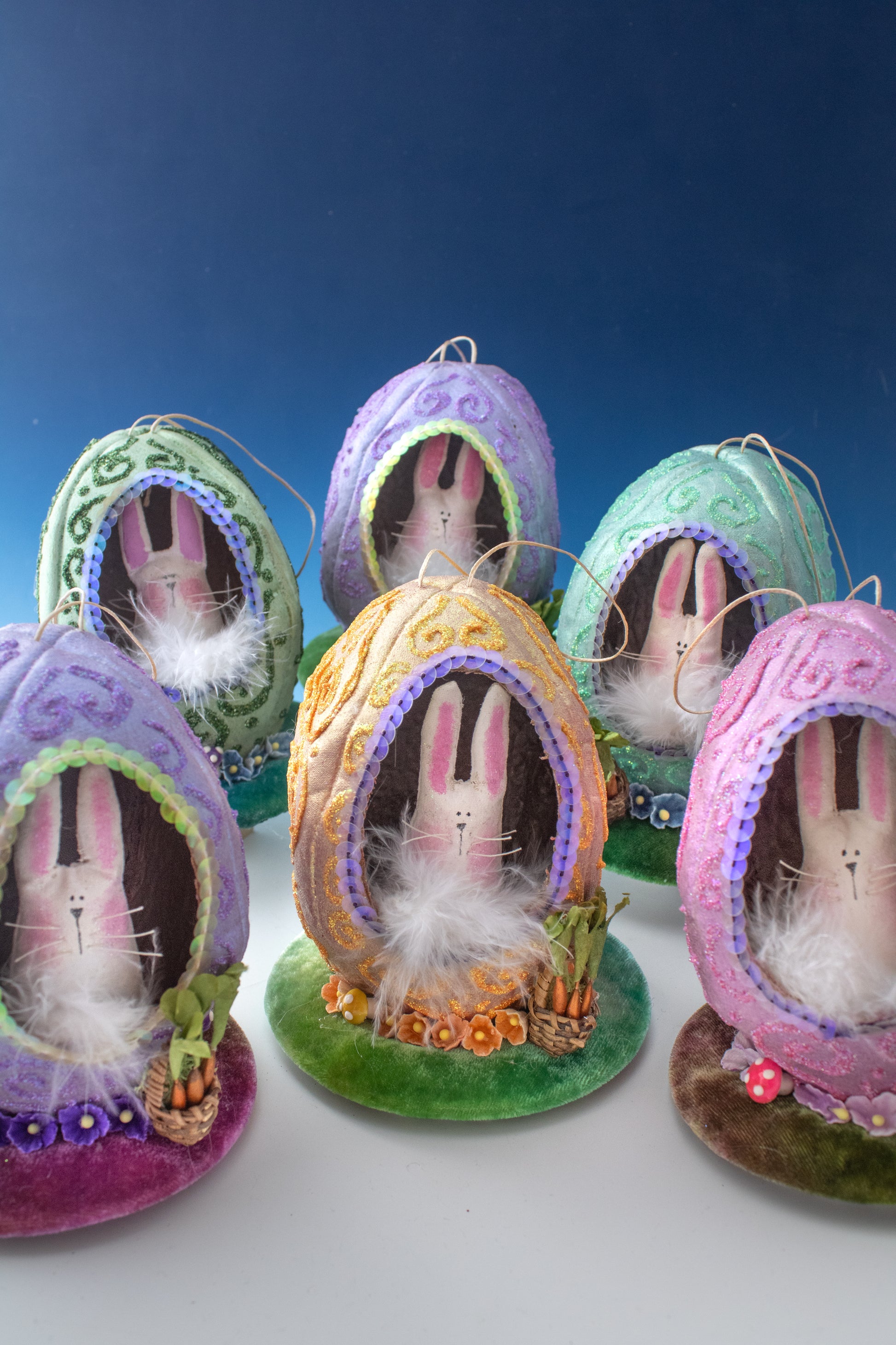 The Creative Spark Easter Egg-stravaganza: DIY Light-Up Diorama features five pastel fabric egg ornaments, each with bunny faces, sequins, faux grass, and twinkling lights, displayed against a blue gradient background.