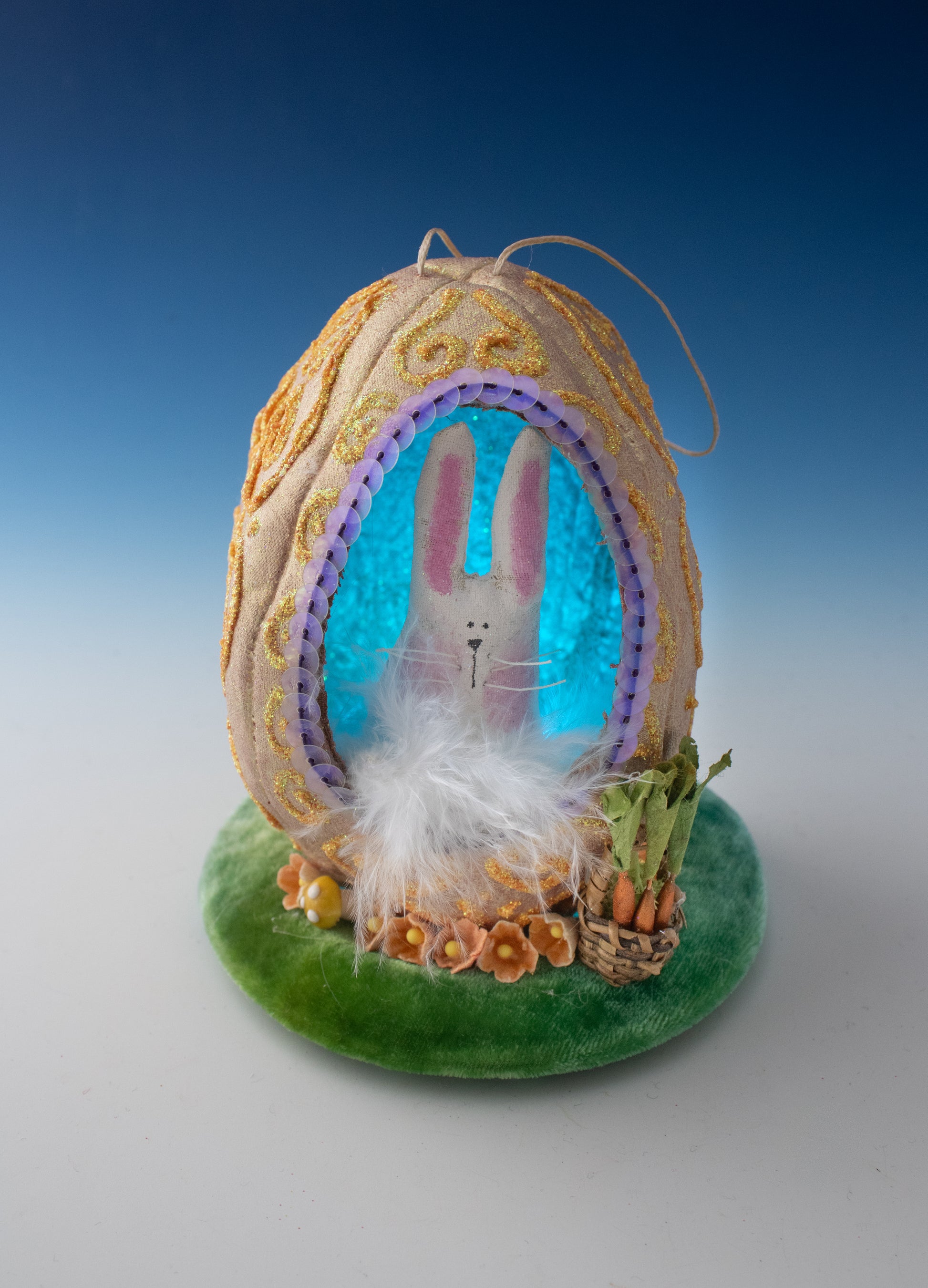 The Creative Spark Easter Egg-stravaganza: DIY Light-Up Diorama features a fabric egg with a cut-out window, revealing a bunny among white feathers. Gold patterns adorn the egg, which sits on a green base with a basket of carrots and flowers.