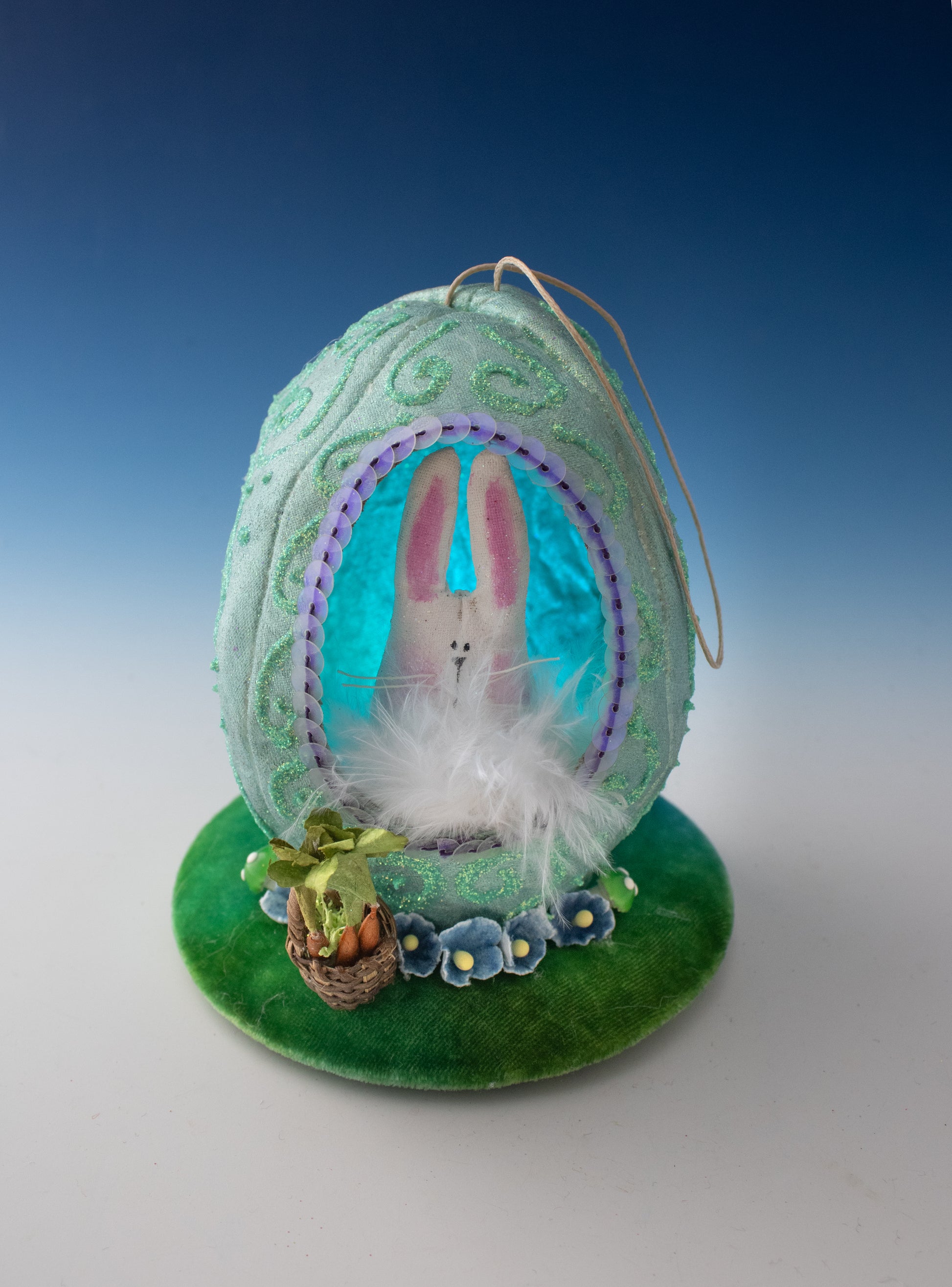 The Easter Egg-stravaganza: DIY Light-Up Diorama by Creative Spark is a pastel blue fabric egg with a cutout showing a white bunny on feathers, set on a green flowered base with plants, and finished with a hanging loop.