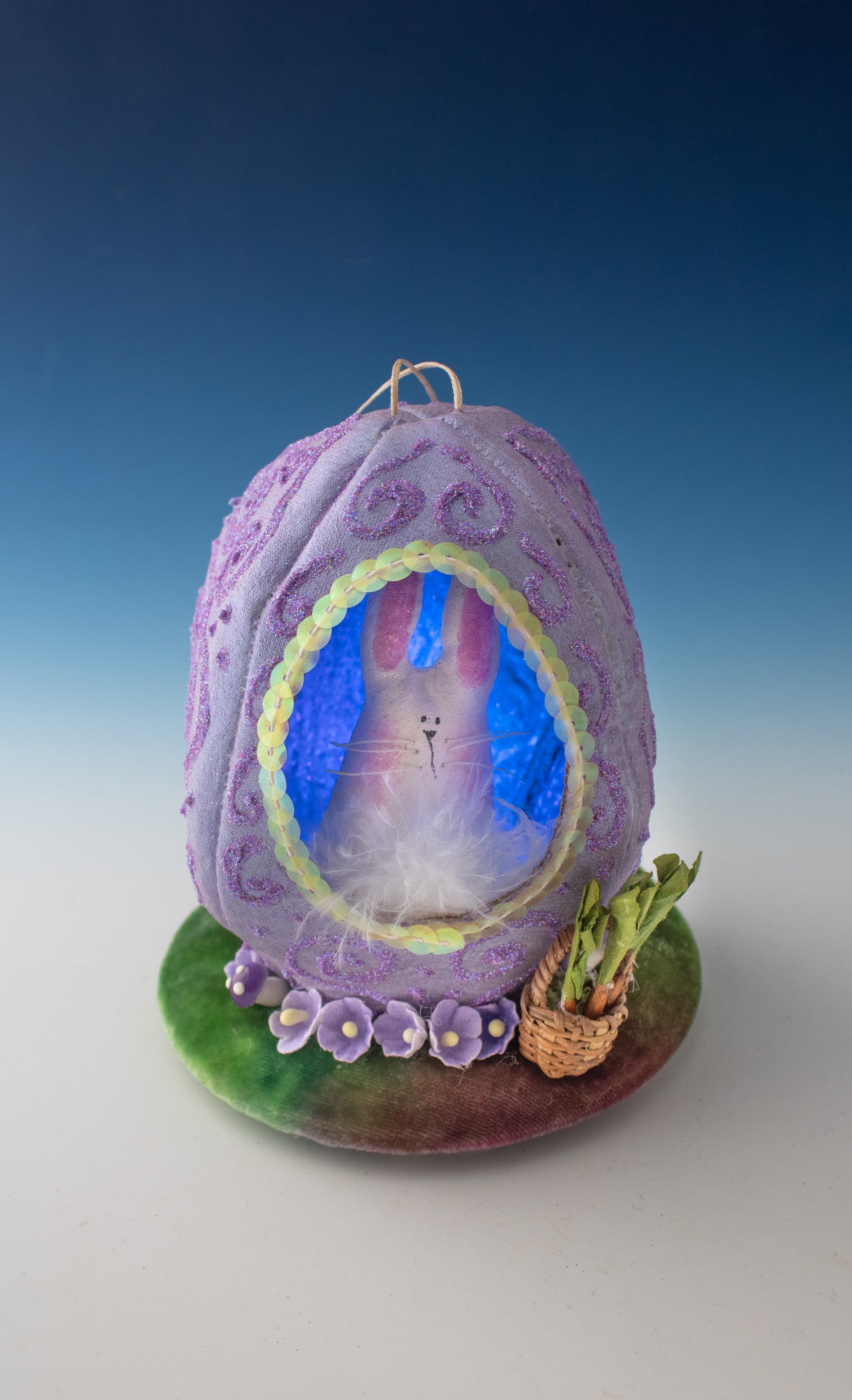 The Creative Spark Easter Egg-stravaganza: DIY Light-Up Diorama features a purple fabric egg with a window displaying a pink bunny inside, set on a green base with purple flowers and a basket of green leaves against a blue-to-white background.