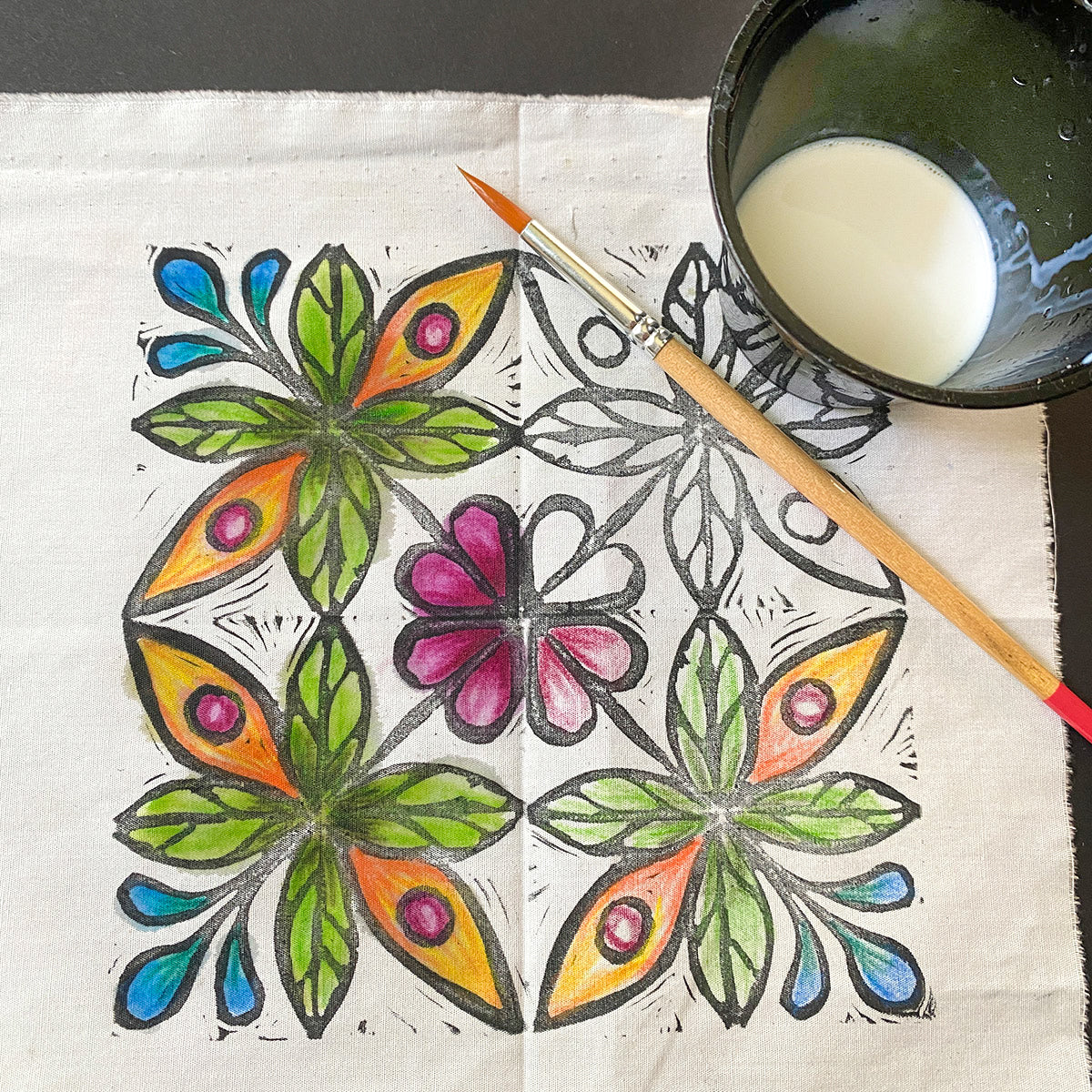 The Coloring Fabric with Inktense Pencils and Pastels by Creative Spark features a painted design of colorful flowers and leaves in blue, green, pink, and orange outlined in black. A paintbrush and a bowl of white liquid on the fabric suggest it was crafted using Inktense pencils.