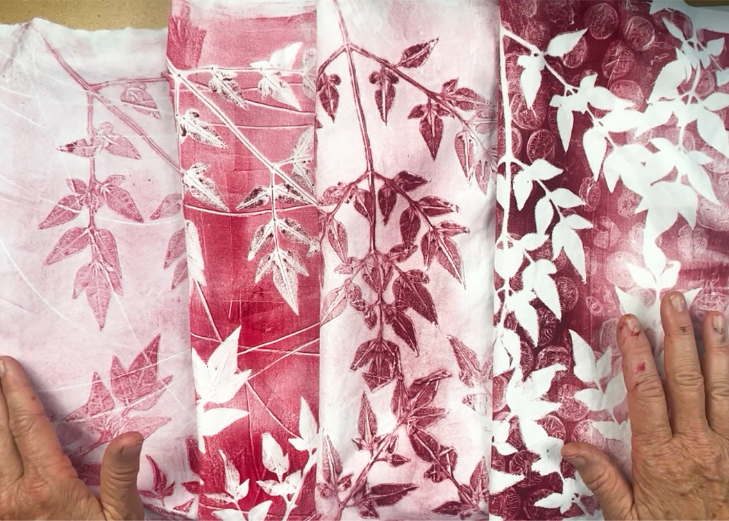 Five papers with red botanical prints, made using the Creative Spark Make Your Own Gel Printing Plate, are displayed side by side. Each sheet shows a unique leaf pattern in red and white as two hands with painted nails hold them at the edges.
