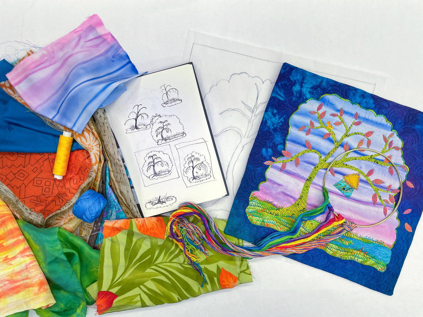 The Creative Spark Wrong Handle Silk Art Quilts Made Simple project features a colorful tree design surrounded by fabric scraps, embroidery floss, yellow thread, blue yarn, and an open sketchbook with tree drawings on a white background.