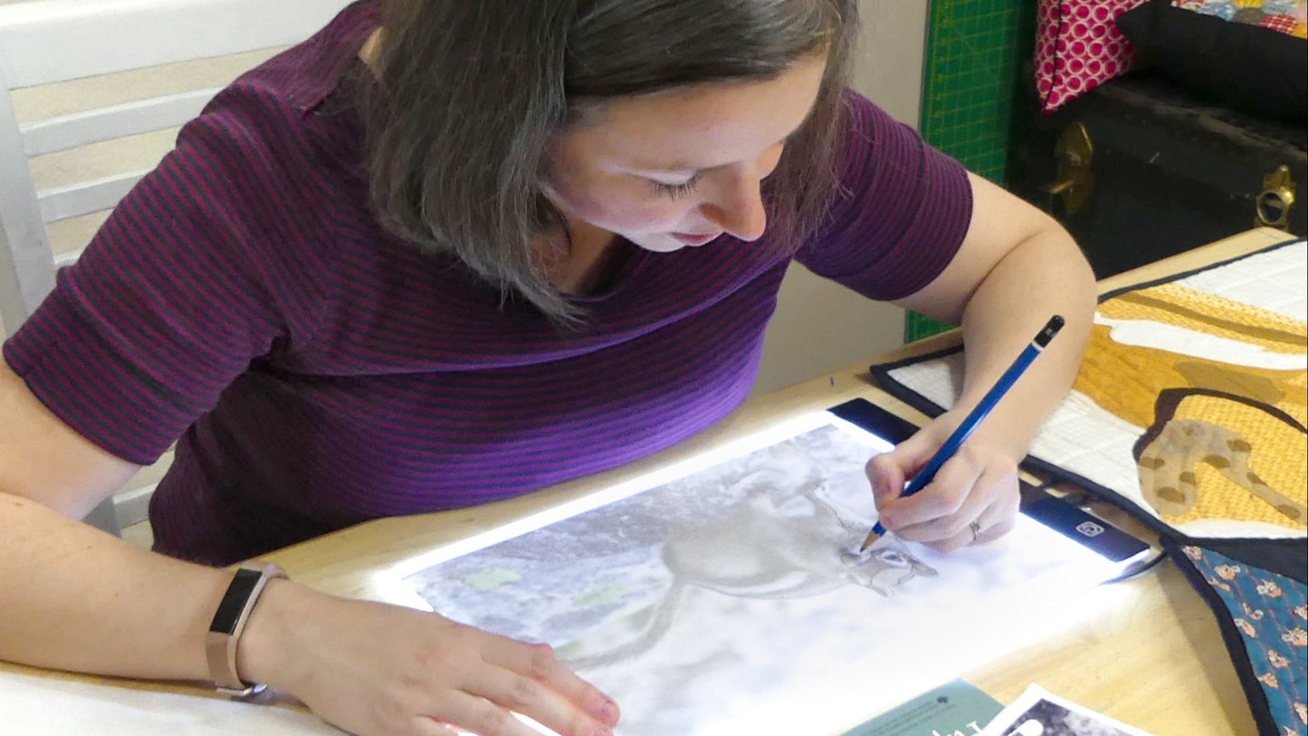 A woman with short brown hair in a purple shirt uses a light box and pencil at a table with art supplies, prepping artwork for the Creative Spark "Design Your Own Paint By Numbers Quilt: Pet Portraits" kit.