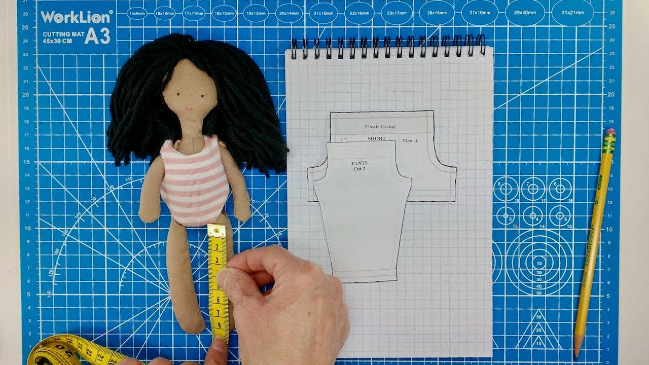 A person uses a measuring tape on a doll's waist next to sewing patterns, set on a blue grid mat—ideal for anyone using Creative Spark’s "How to Alter Doll Clothes Patterns" for custom designs.