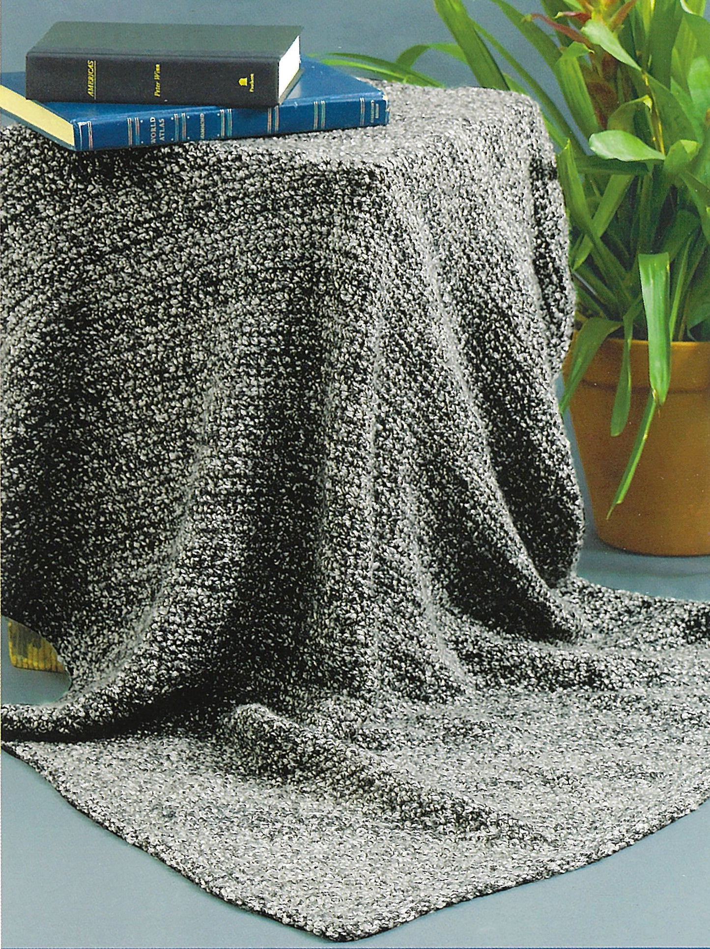 A gray textured blanket covers a round table. Three stacked books—including the "Learn To Knit—Lion Brand Yarn Booklet" by Leisure Arts—invite you to knit, while a green potted plant sits on the floor next to the table.