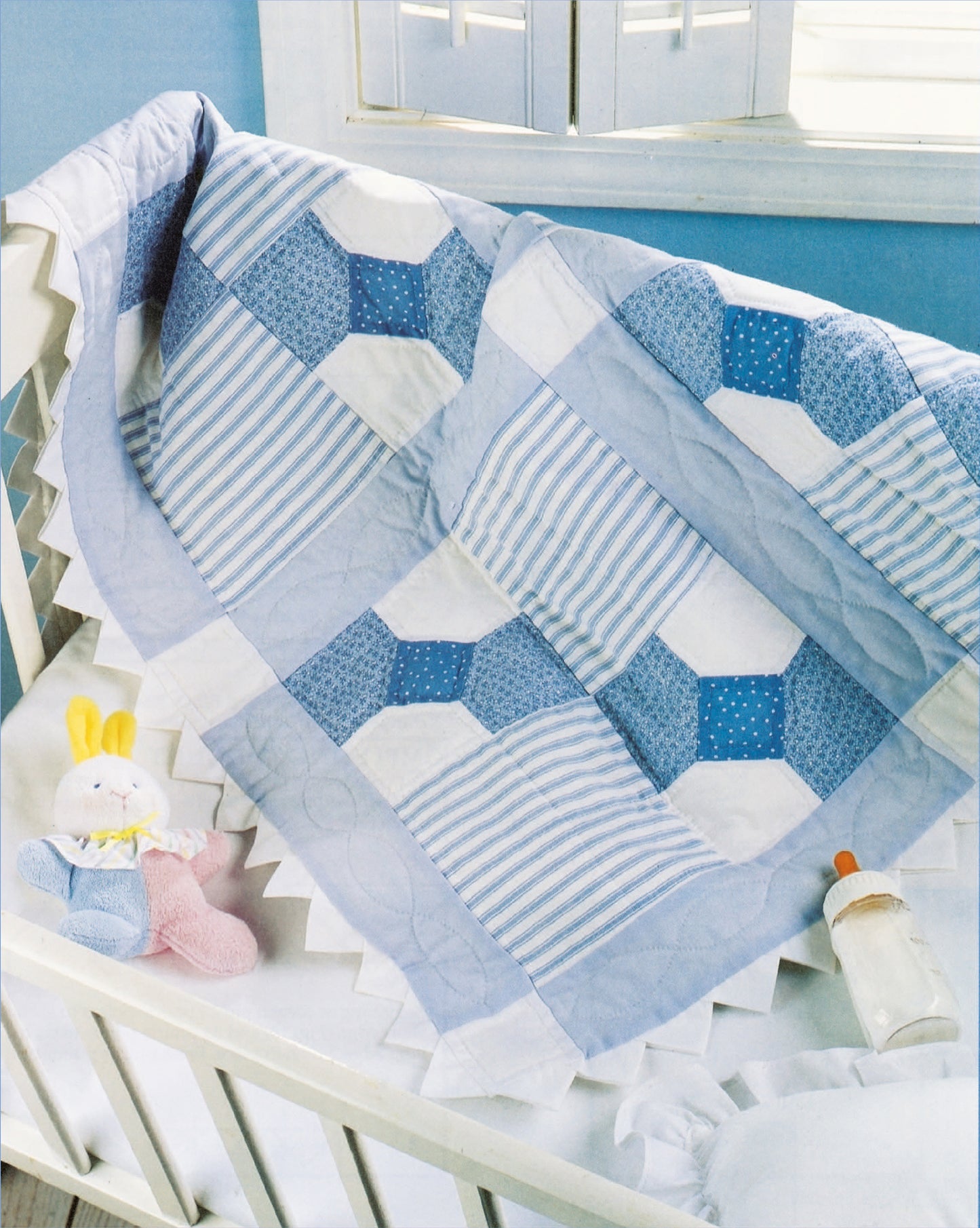 The Rock-a-Bye Quilts by Leisure Arts features a blue and white nursery rhyme quilt, a stuffed bunny, pink rattle, and baby bottle in a crib, set by a sunlit window against a light blue wall.