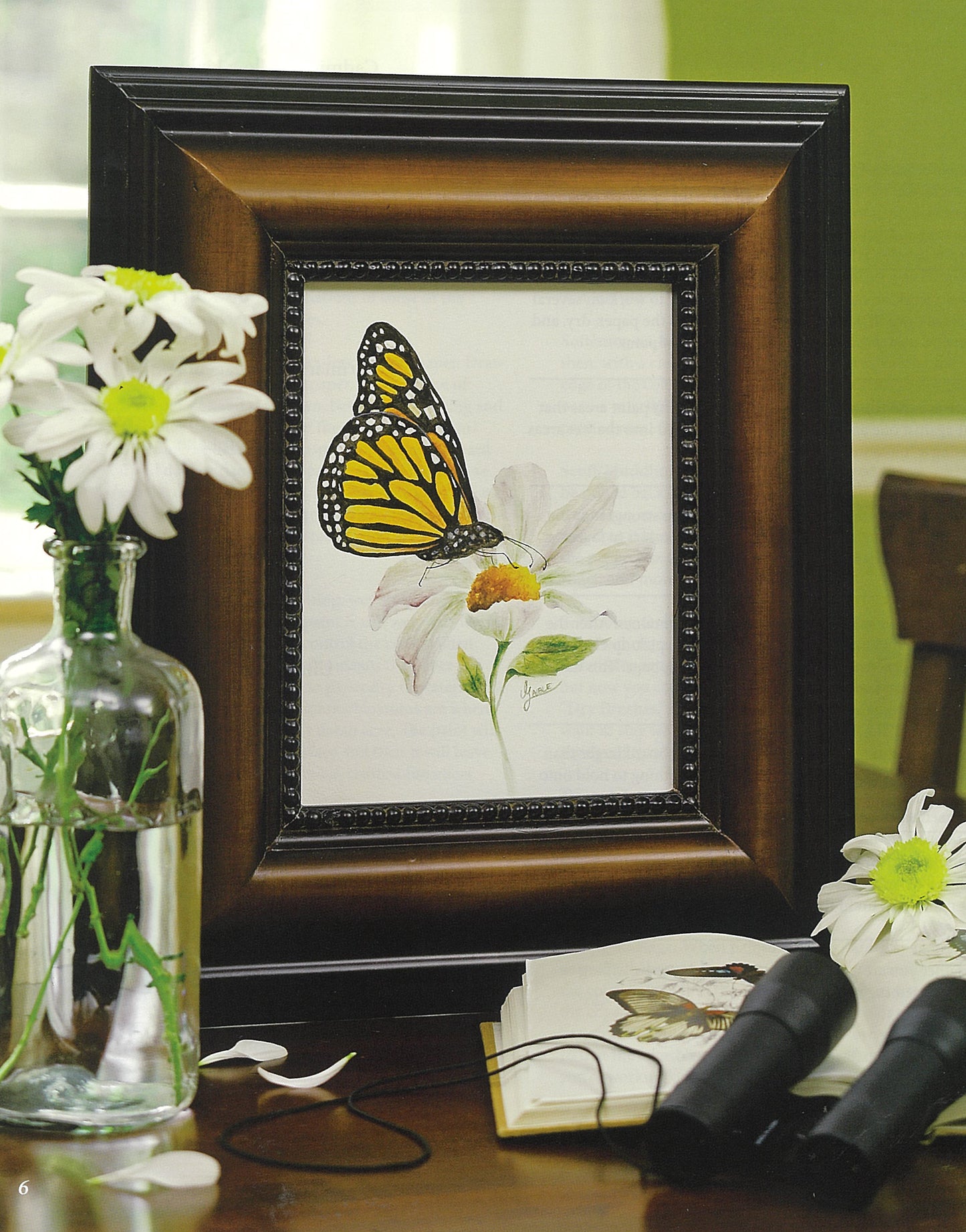 A Deborah A. Lambein framed painting of a monarch butterfly on a daisy sits near daisies in a glass, a book, and binoculars. The sunlit green background from Leisure Arts’ Watercolor Classics—7 Beginner Friendly Designs offers peaceful inspiration.