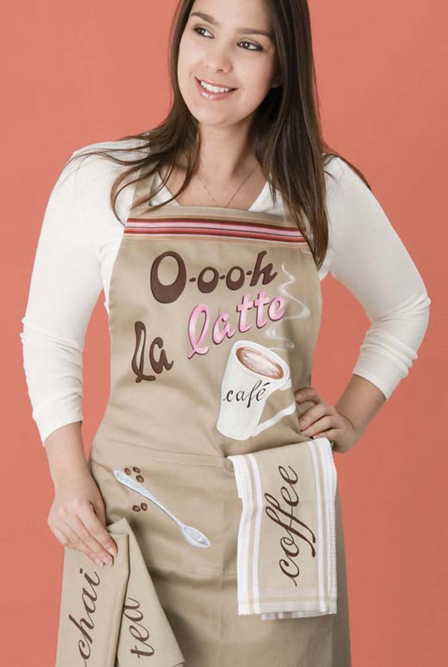 A woman stands against a coral background wearing The Painted Kitchen apron by Leisure Arts, holding a chai tea towel and a striped coffee towel—stylish kitchen decor perfect for DIY cooking projects.