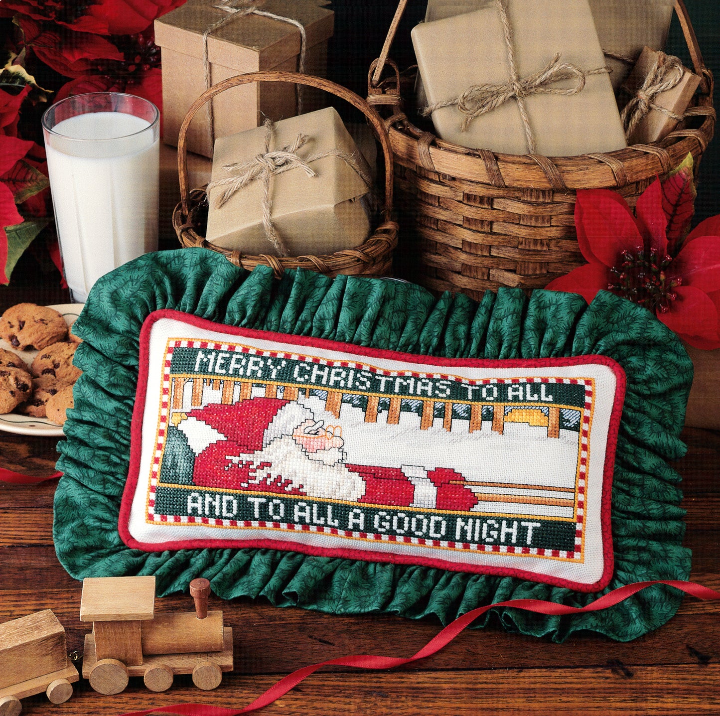 A festive scene with wrapped gifts, cookies, milk, and a pillow inspired by Santa's Great Book of Cross Stitch from Leisure Arts featuring a Christmas Santa design and “Merry Christmas to all and to all a good night.”.