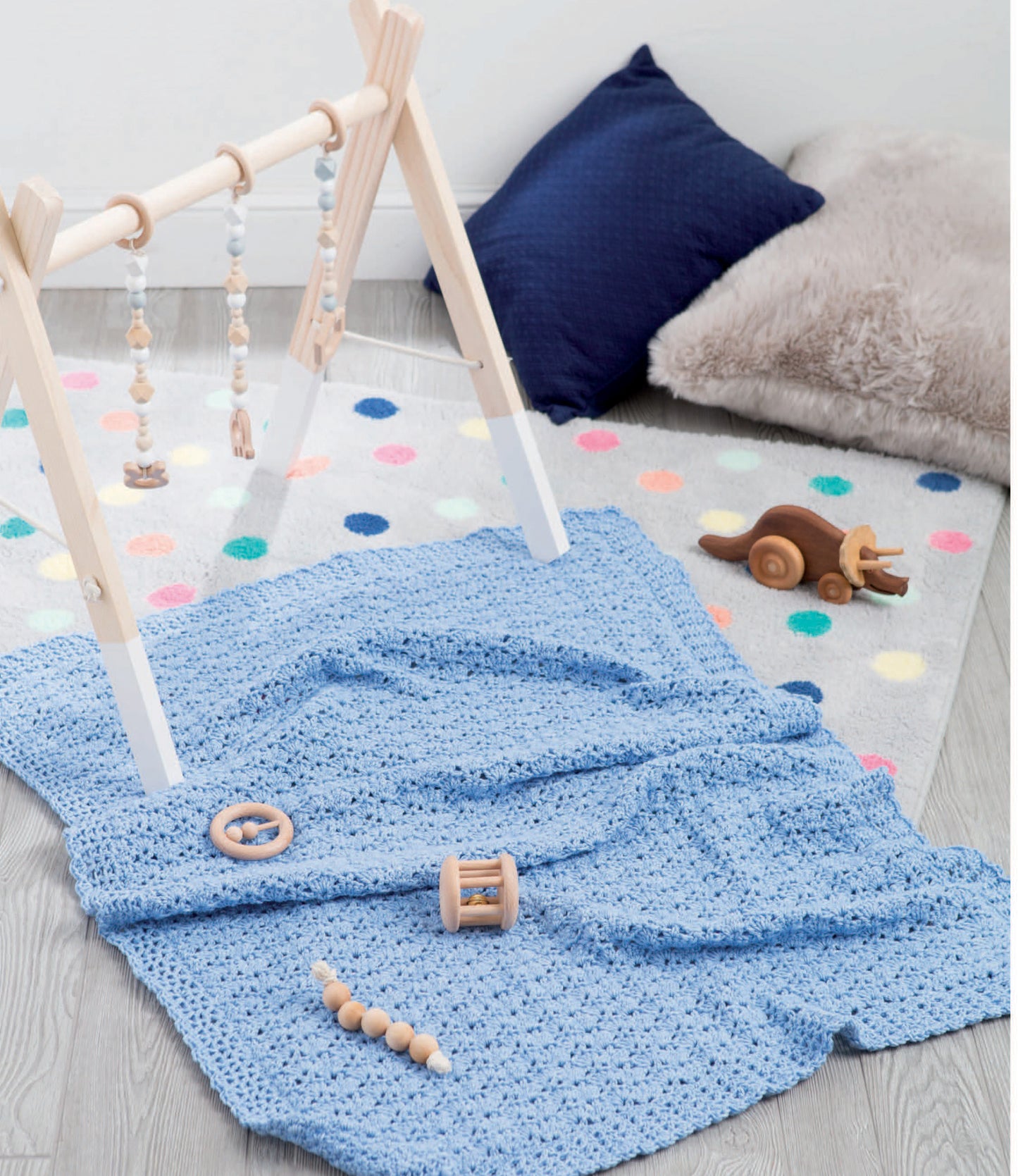 Create a cozy play area with "Our Best Baby Afghans" by Leisure Arts in blue, a wooden baby gym, plush gray and dark blue pillows, a colorful dotted rug, and wooden toys on a light wood floor—perfect for beginner crochet enthusiasts.