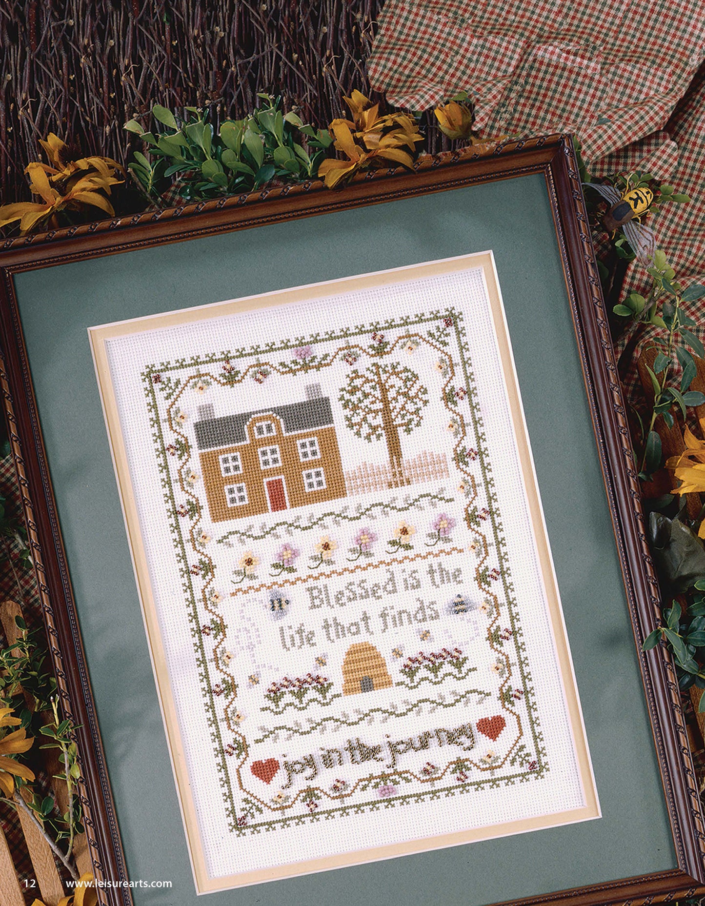 The Joy in the Journey Digital Download by Leisure Arts features a cross-stitch house, tree, flowers, bees, and beehive with heart and floral borders. It reads: Blessed is the life that finds joy in the journey. Greenery surrounds the frame.