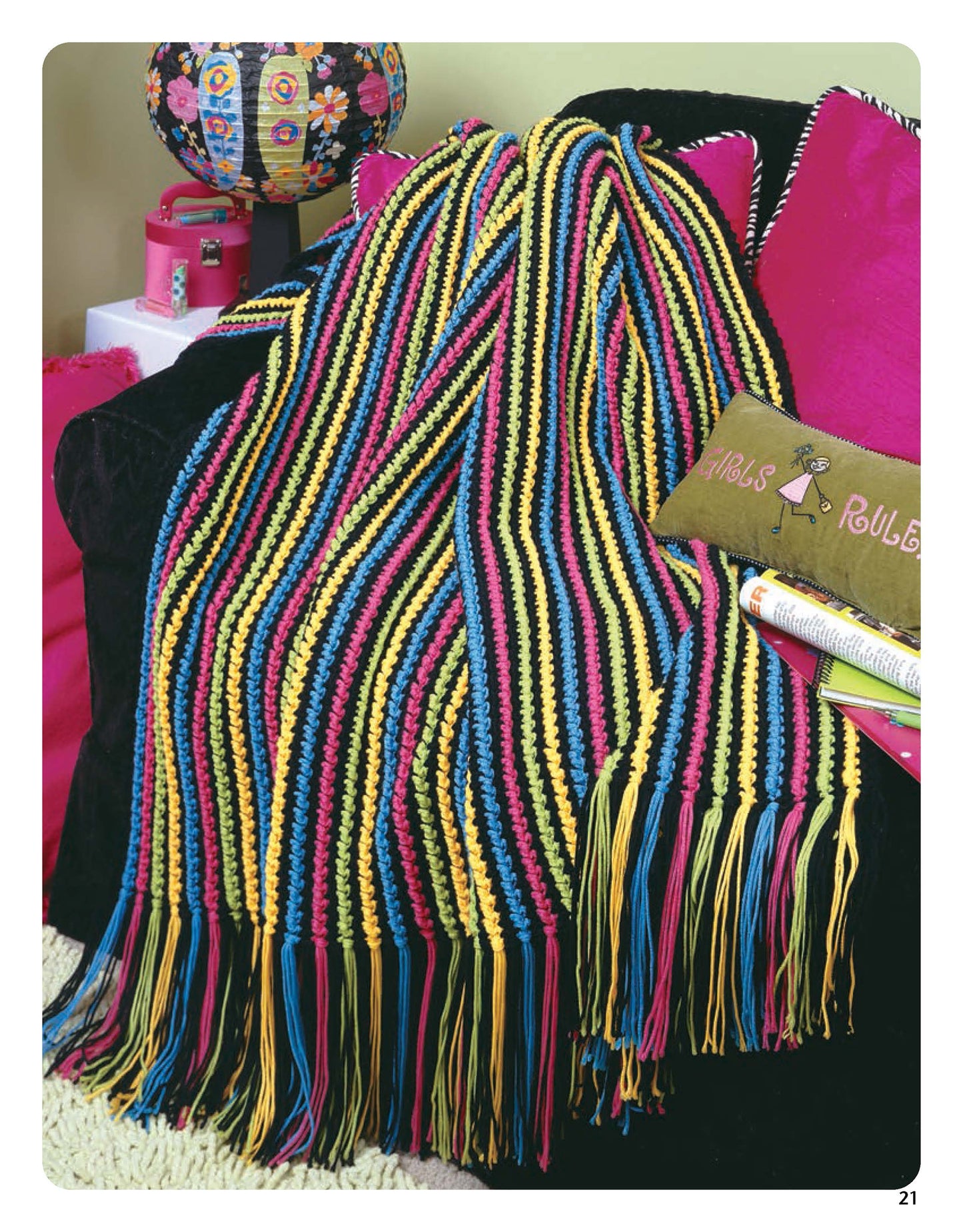 The "Color-Charged Throws" by Leisure Arts features yellow, blue, pink, and black striped crochet with long tassels, draped over a black sofa with bright pillows—a vibrant and easy project inspired by Leisure Arts designs.