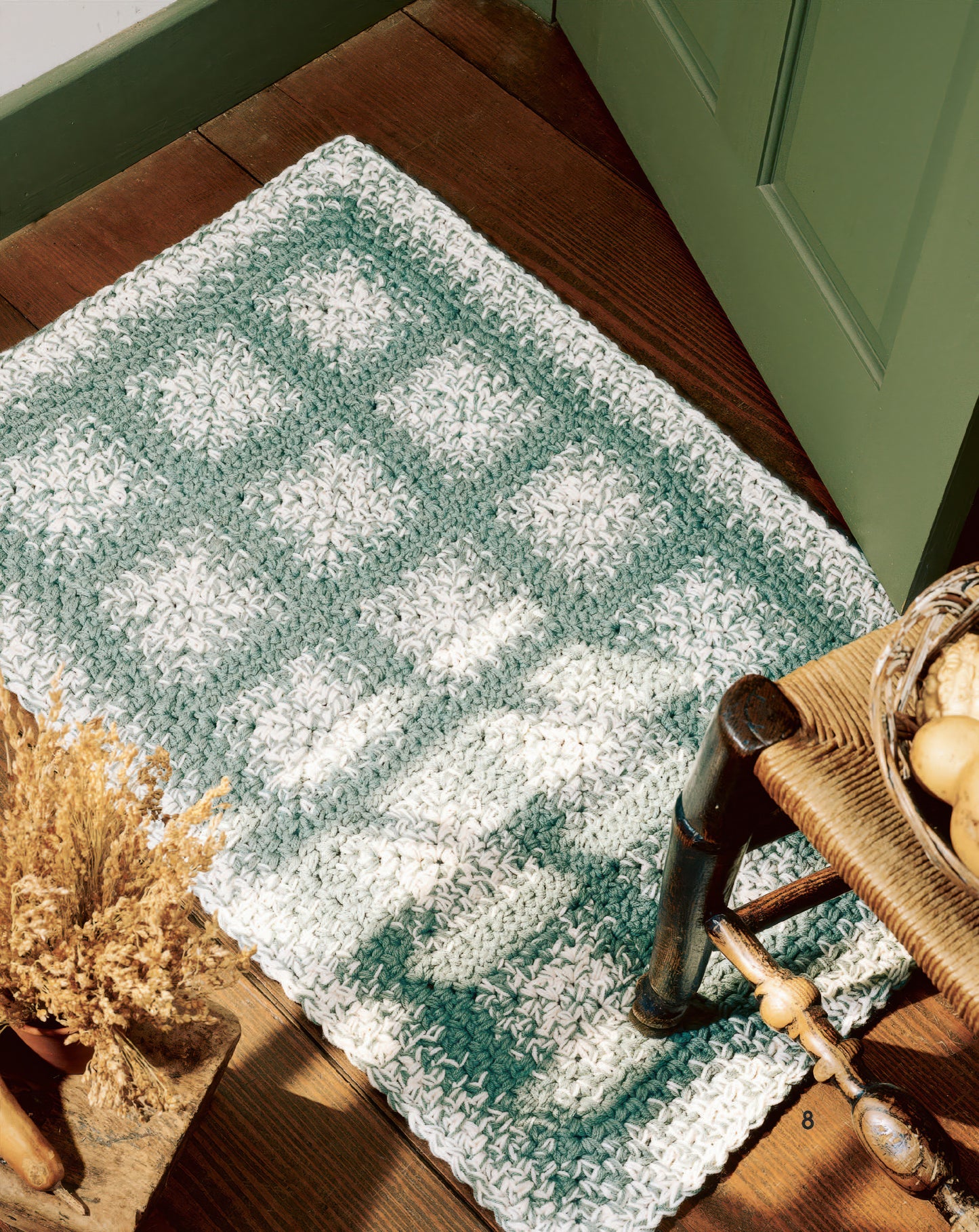 The "Easy Rugs to Crochet" digital download by Leisure Arts features a green and white checkered rug, ideal for beginners, displayed on a wooden floor near a green door with a chair and dried flowers in view.