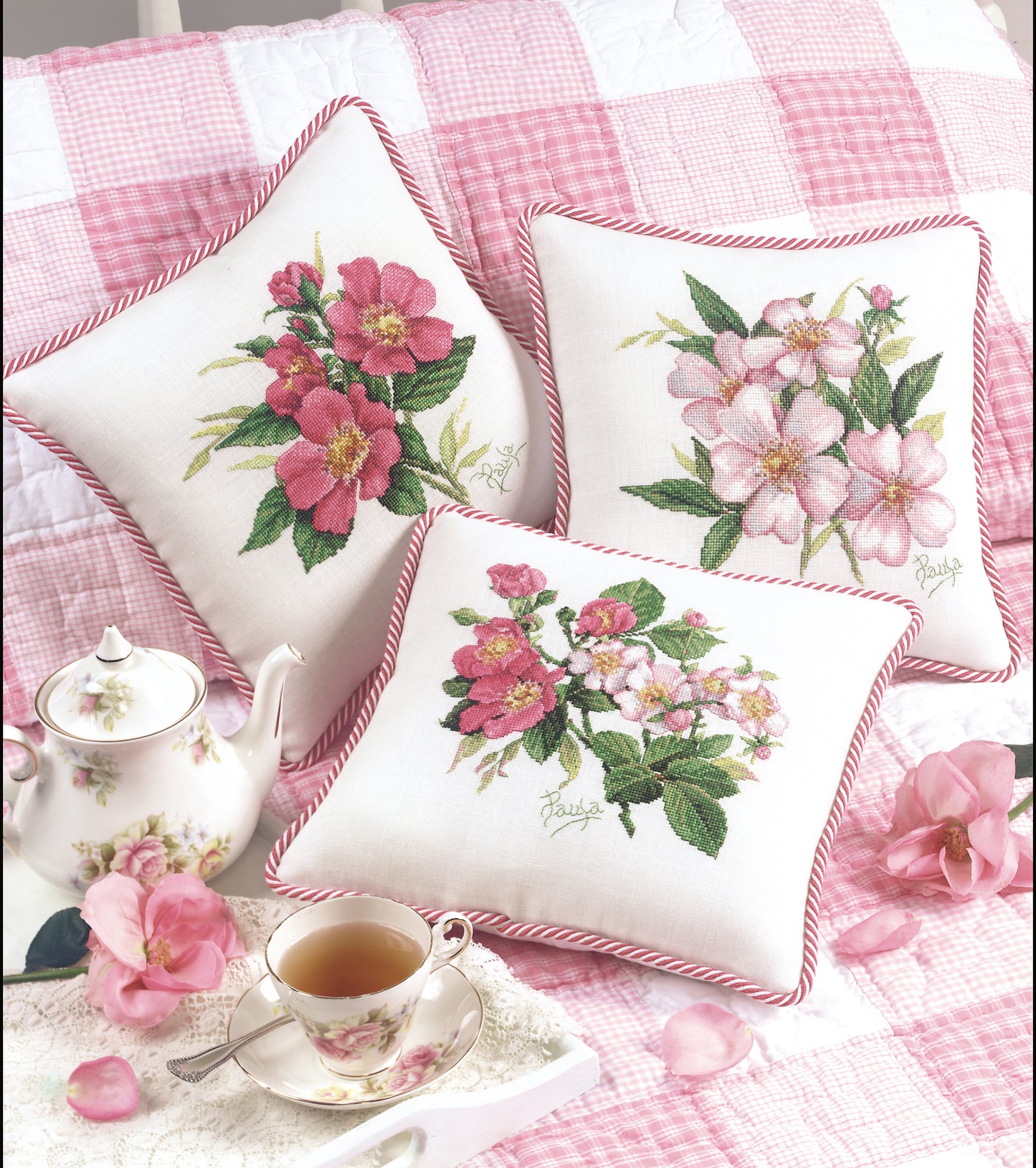 Three pillows from Paula's Garden Digital Download by Leisure Arts, featuring white fabric with pink flower embroidery, rest on a pink and white checkered quilt beside floral teaware and scattered pink petals.