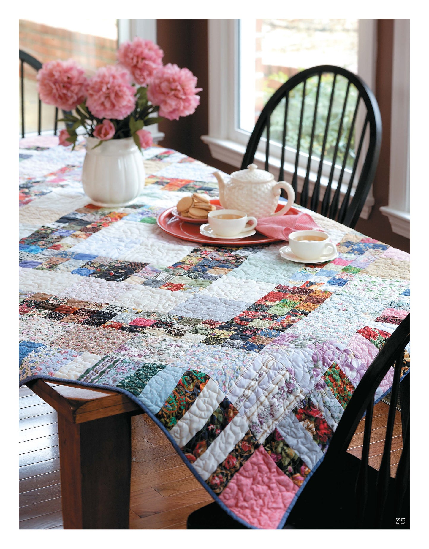 A vibrant patchwork quilt made with quick piecing from Leisure Arts' Slash Your Stash Quilts Digital Download graces a wooden table dressed with a white teapot, teacups, macarons, pink flowers, black chairs, and large windows.