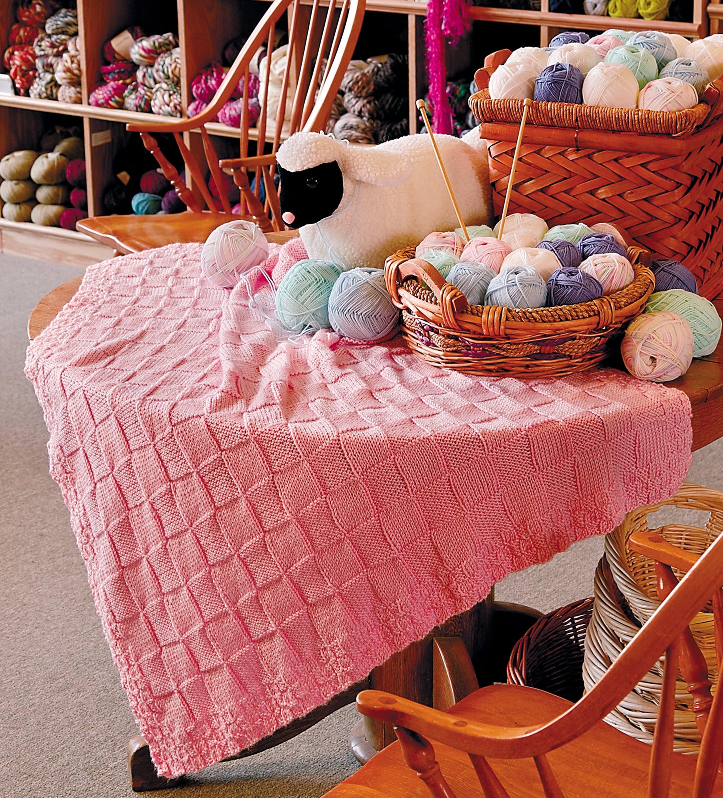 A pink knitted blanket drapes over a table in a cozy yarn shop, surrounded by colorful yarn and needles—perfect inspiration for crafting Leisure Arts’ Knit Along with Debbie Macomber—The Shop on Blossom Street.