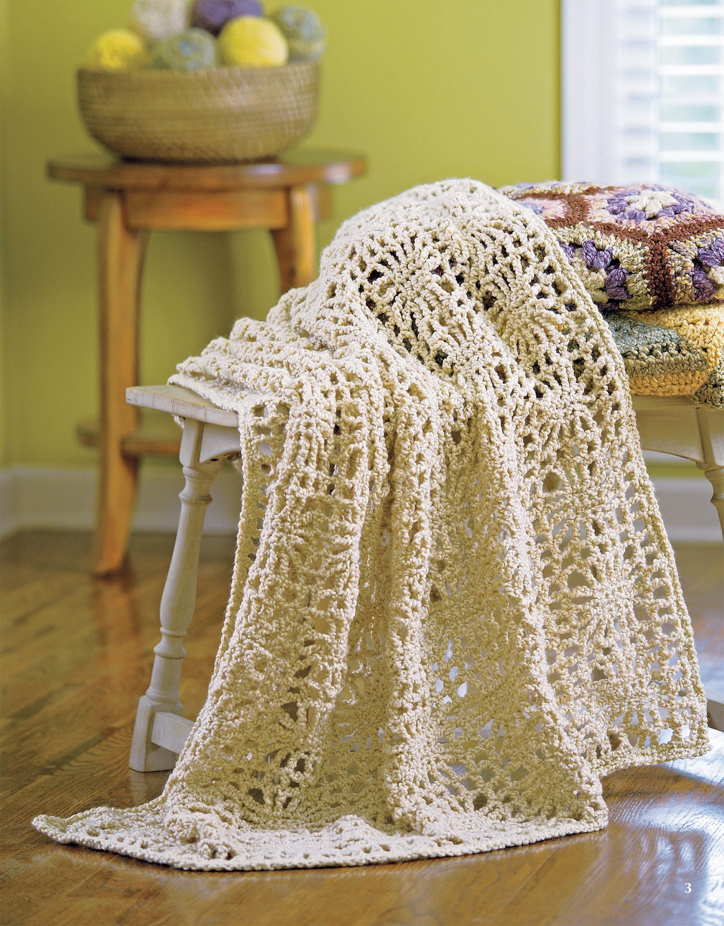 A cream-colored crocheted blanket from the Homespun Afghans collection by Leisure Arts drapes over a white wooden bench. Nearby, folded quilts and a basket of yarn create a cozy vibe in the sunlit room with light green walls.