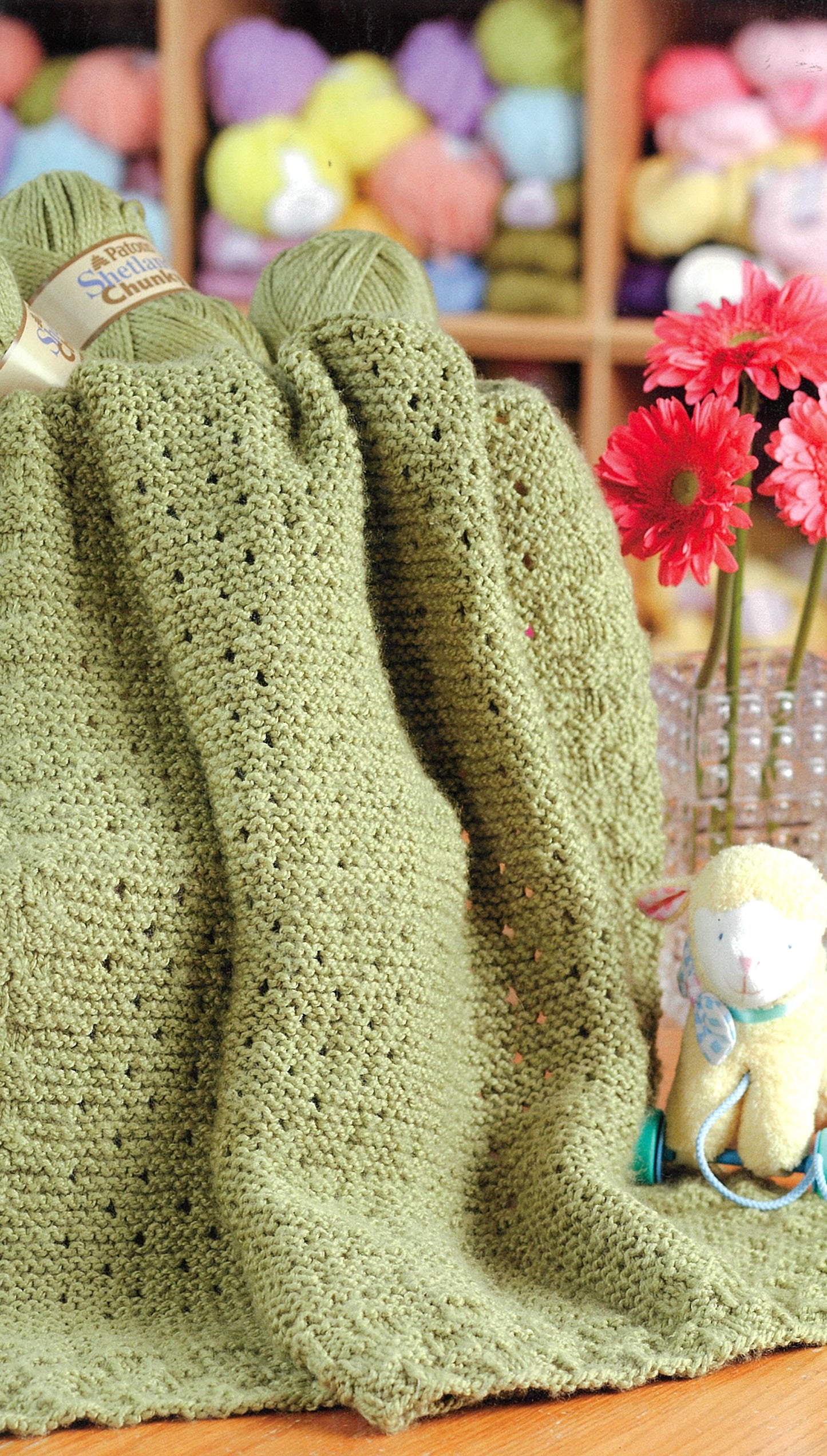A green knitted blanket drapes over a table with yarn balls and red flowers nearby. A stuffed animal sits by shelves of colorful yarn—inspiration for Leisure Arts’ Knit Along with Debbie Macomber—Back on Blossom Street.