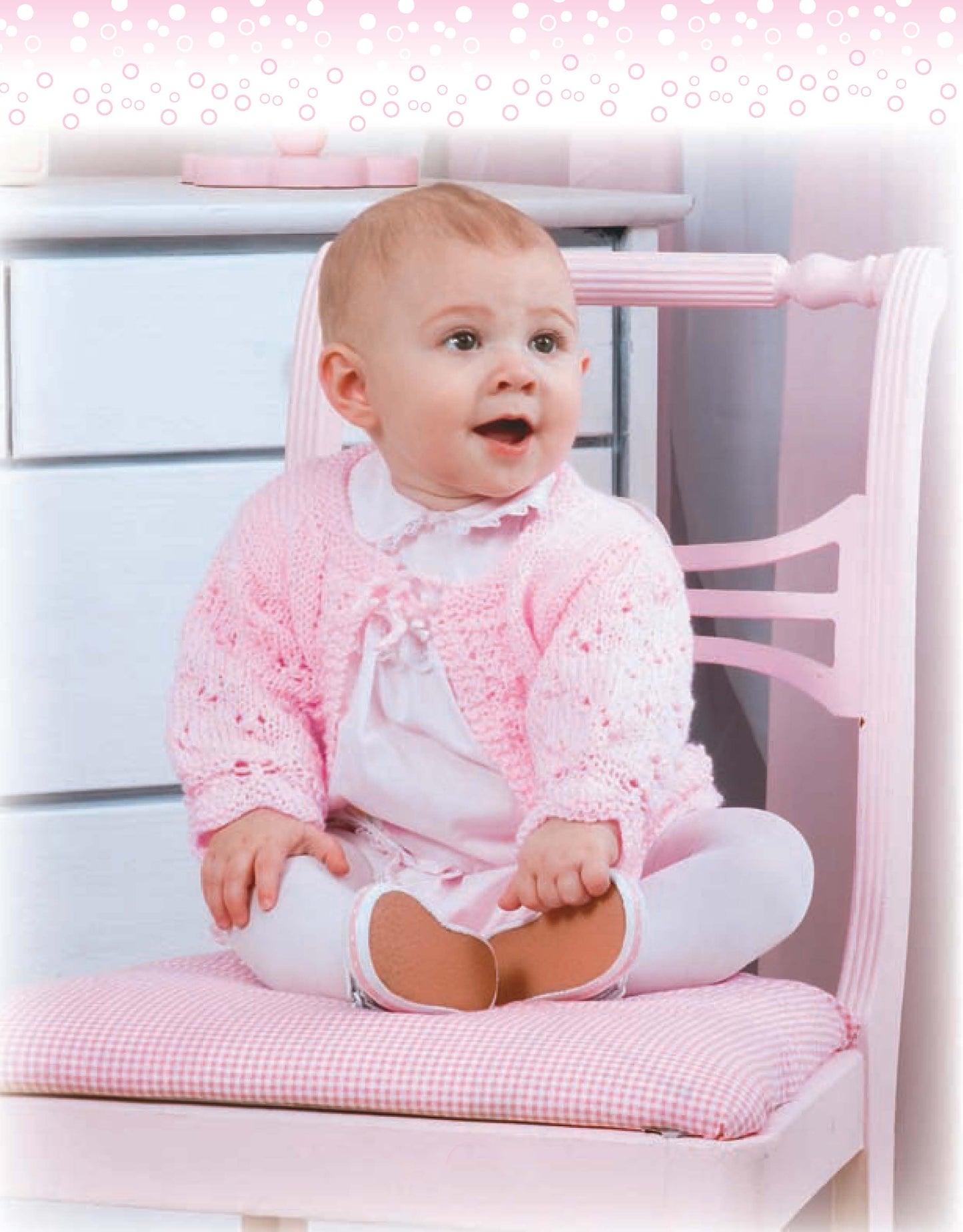A baby in a pink sweater from Leisure Arts’ Lacy Baby Sets to Knit, with white tights and shoes, sits smiling on a pink chair with checkered cushion. A dresser and a dotted border appear in the background.