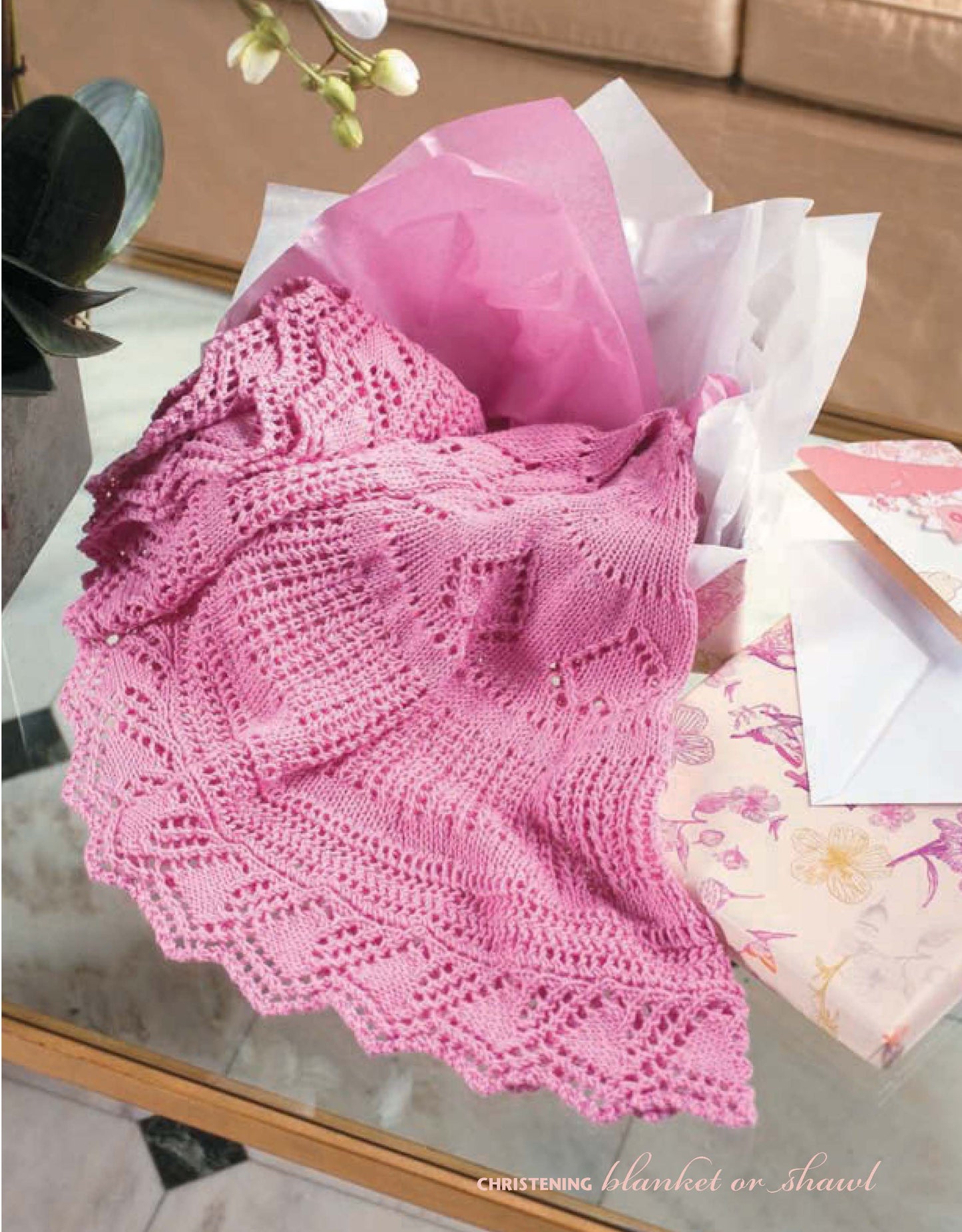 A pink lace knit piece from Leisure Arts' "I Can't Believe I'm Lace Knitting" is draped over tissue paper beside a floral greeting card on a marble table, with an orchid and beige sofa in the background.