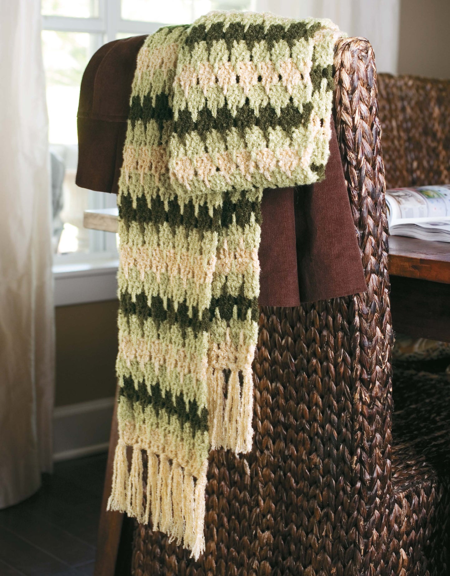 The "Learn a Stitch Create a Scarf Digital Download" by Leisure Arts features a crocheted chevron scarf in green, cream, and brown, finished with fringe and perfect for cozy projects—displayed on a woven chair by a window.