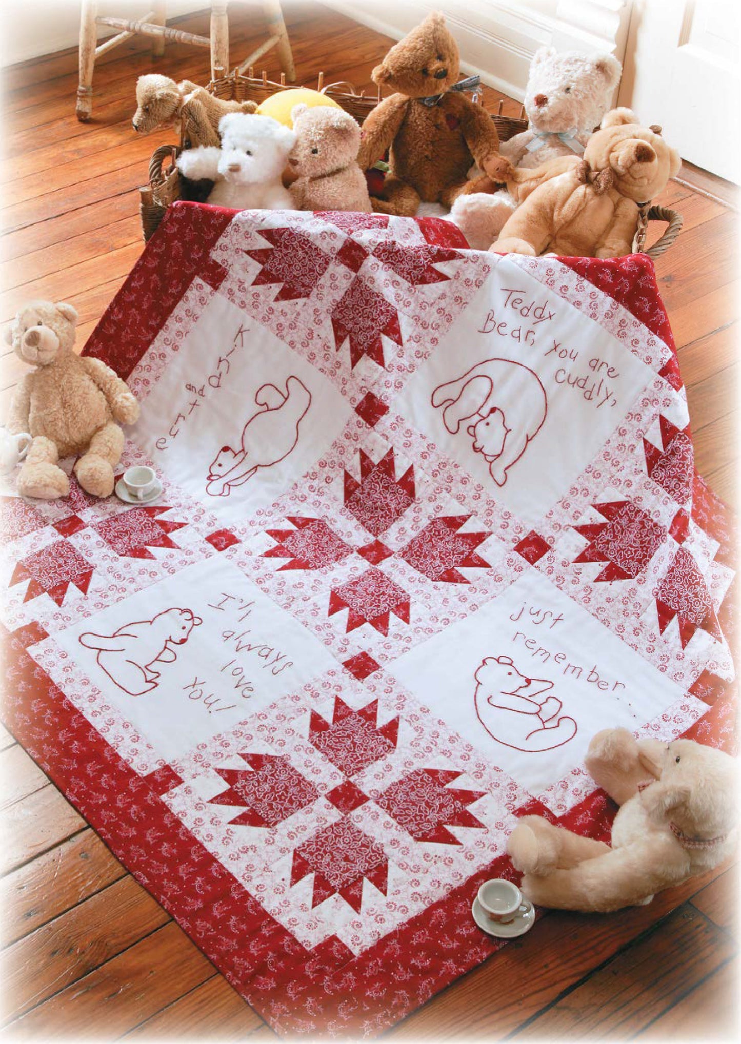 The Quilt Along with Emilie Richards by Leisure Arts, a red and white patchwork quilt with embroidered teddy bears and messages, is displayed on a wooden floor among teddy bears and two toy tea cups with saucers.