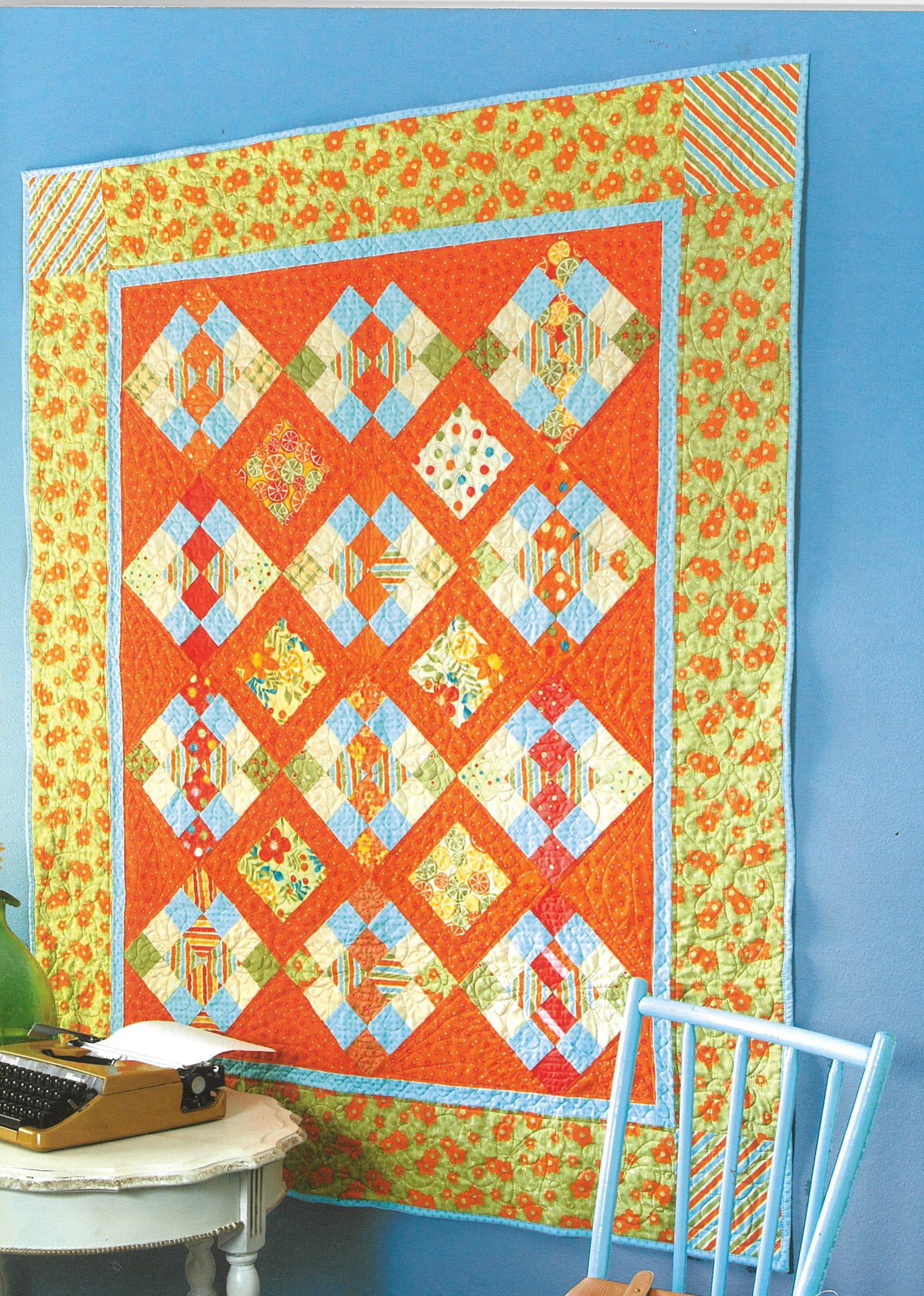 Pat Sloan's Camera, Color, Quilts! by Leisure Arts features a vibrant patchwork quilt with orange, yellow, blue, and green diamond patterns hanging on a blue wall near a white table with a typewriter and a light blue chair.