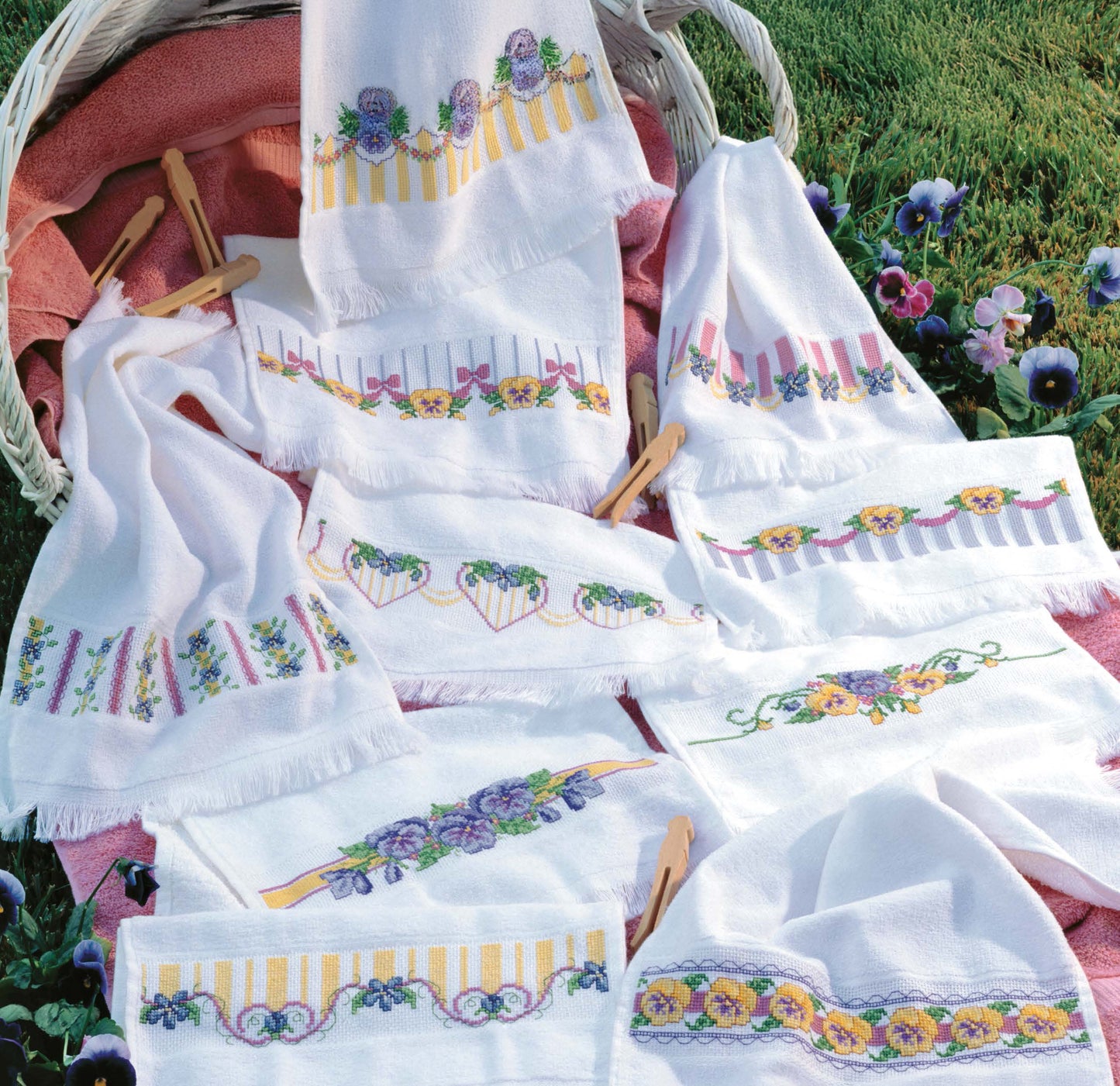 Spring Florals For Fingertips by Leisure Arts features white hand towels with colorful embroidered flowers and stripes, displayed on grass with a basket, clothespins, and purple blooms—ideal for charming home decor.