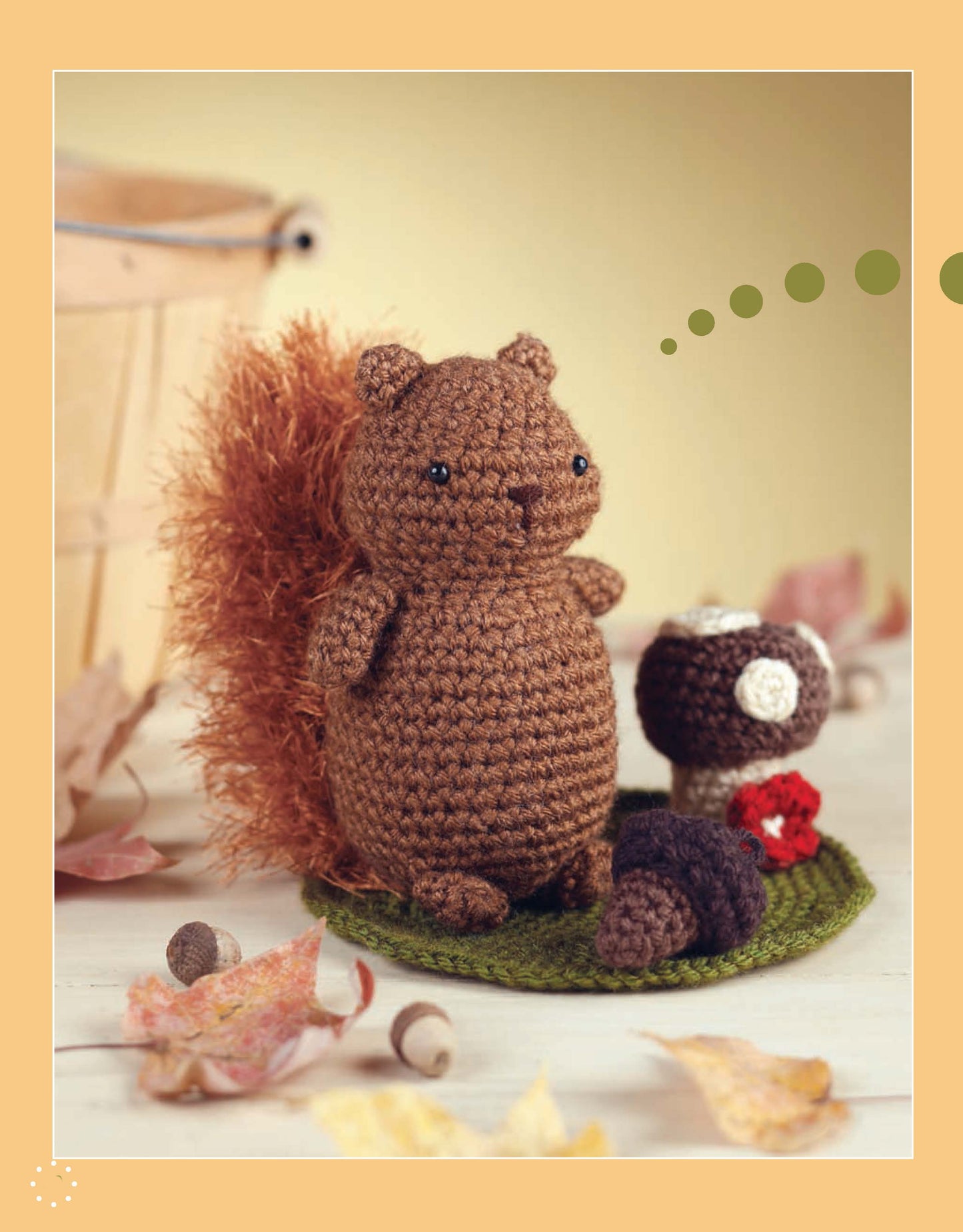 A crocheted brown squirrel with a fluffy tail stands on a green mat surrounded by mushroom, ladybug, and acorn toys from the Leisure Arts book "More Cute Little Animals To Crochet"; a wooden basket is in the background.