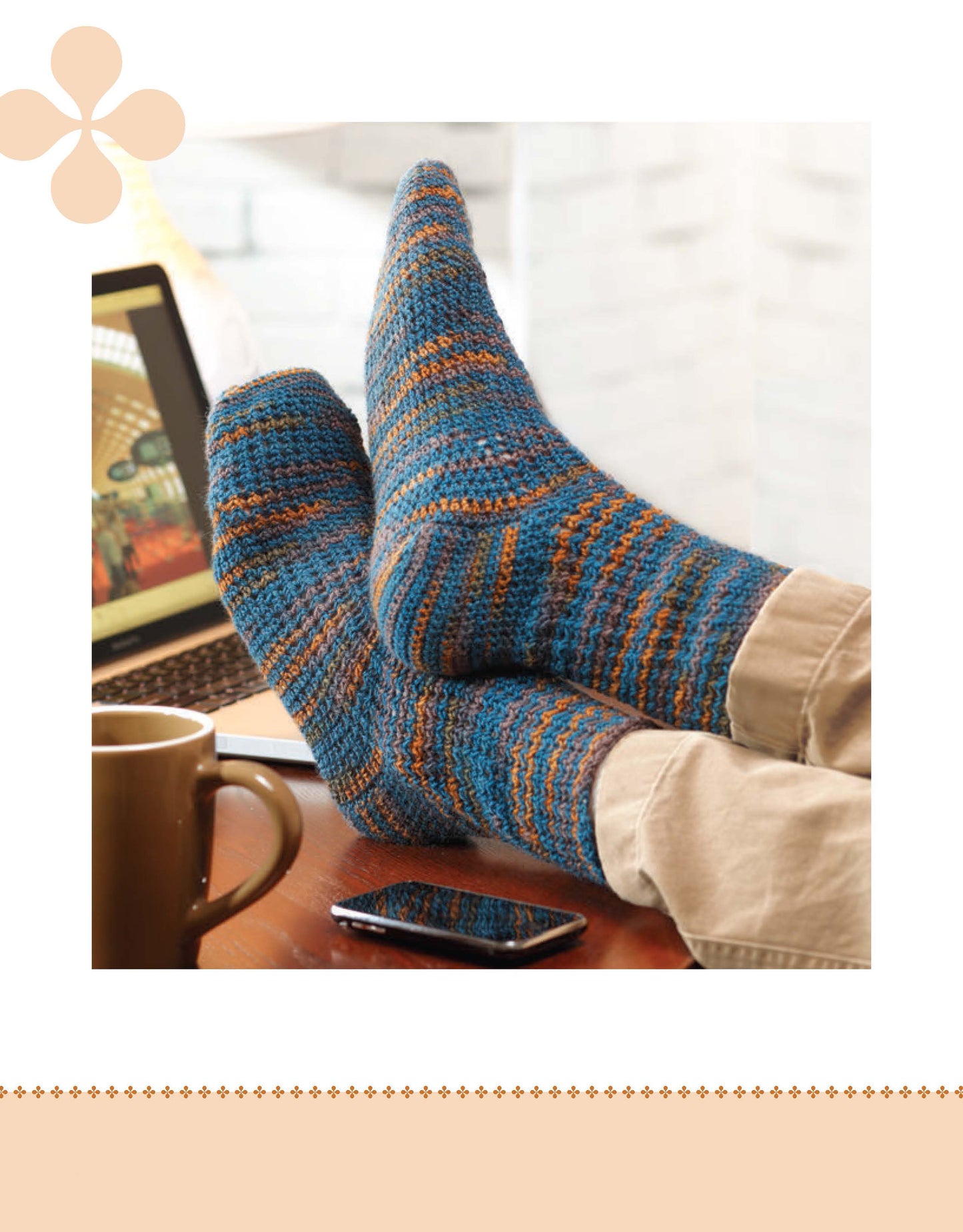 A person wearing blue and orange crochet socks relaxes with a mug, smartphone, and laptop—an ideal moment to browse Leisure Arts' “I Can't Believe I'm Crocheting Socks” digital download for beginner-friendly crochet patterns.