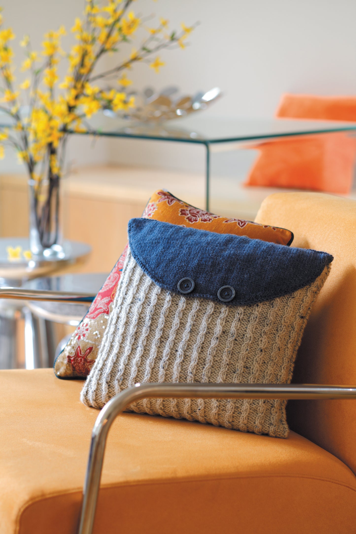 A knitted pillow from "The Envelope, Please!—Ten Knit Designs" by Leisure Arts, made with worsted weight yarn and blue flap with buttons, sits on an orange chair. A floral cushion and glass table with yellow flowers are in the background.