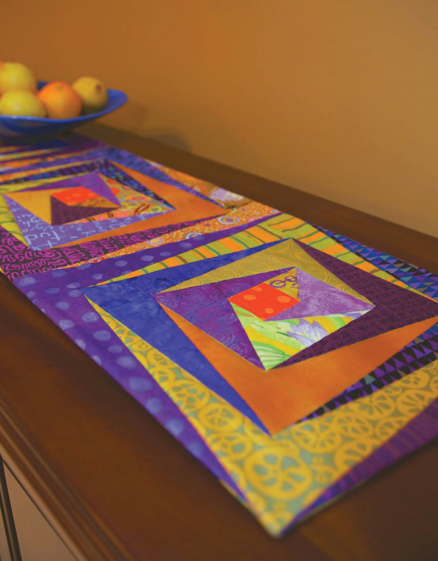 The "Quilt Blocks Go Wild!" table runner from Leisure Arts features vibrant geometric patterns in purple, yellow, orange, and green. Displayed on wood with a blue apple bowl in the background, it offers great inspiration for beginner quilters.