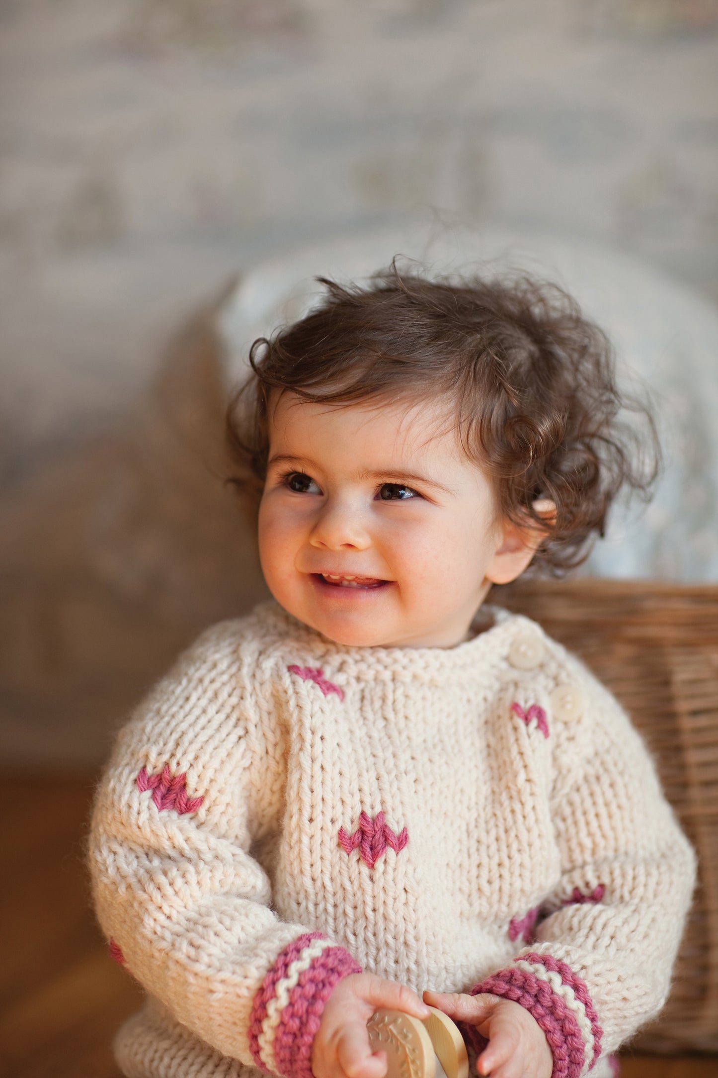 A smiling toddler with curly brown hair wears a cream sweater featuring pink baby patterns from Leisure Arts' "Knit in a Day for Baby" as they sit indoors holding a round object, with the background softly blurred.