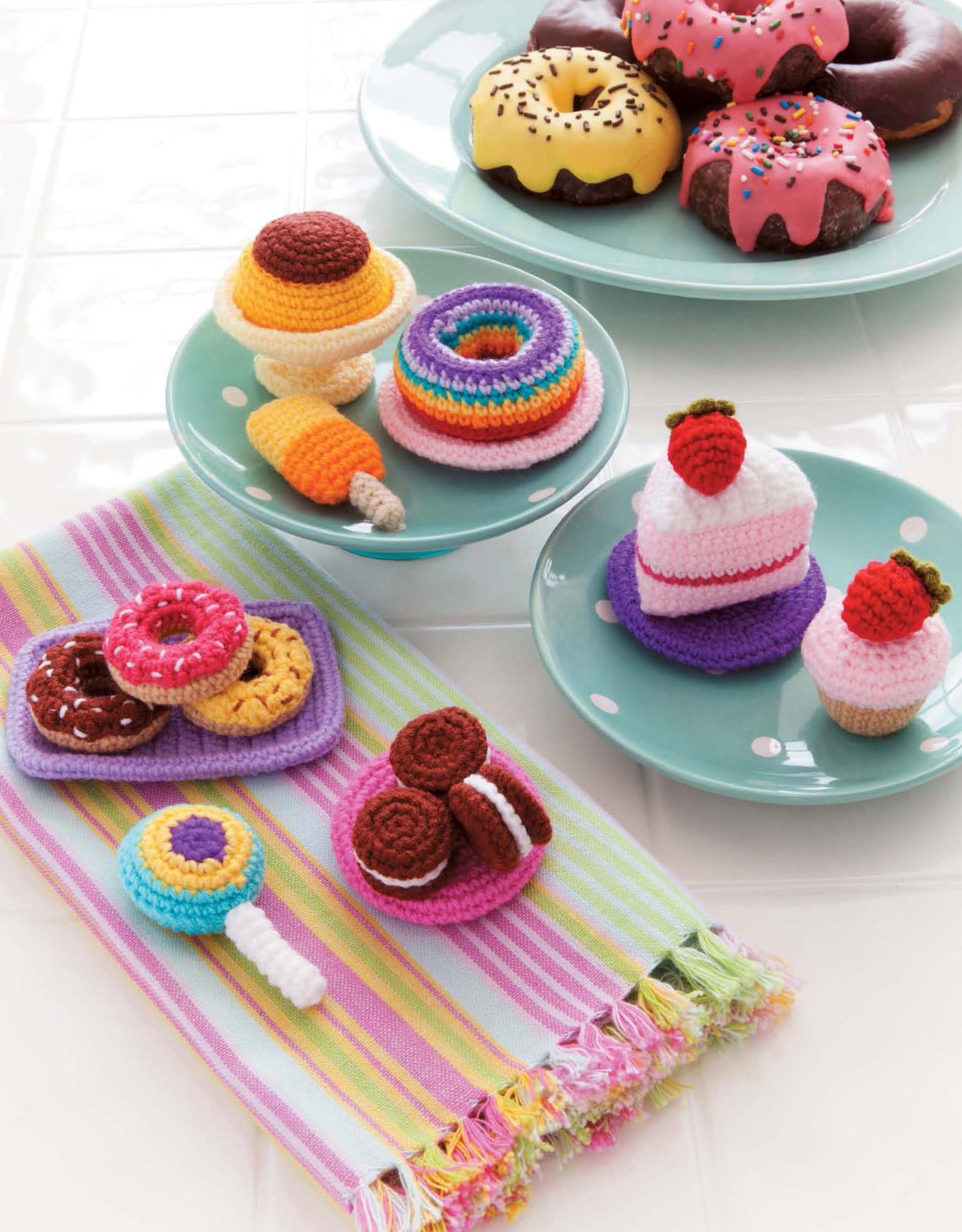 Colorful crocheted desserts from the "Ice Box Crochet" collection by Leisure Arts, including donuts, cakes, cookies, and lollipops, are displayed on pastel plates and a striped cloth, mimicking real sweets on a white tiled surface.