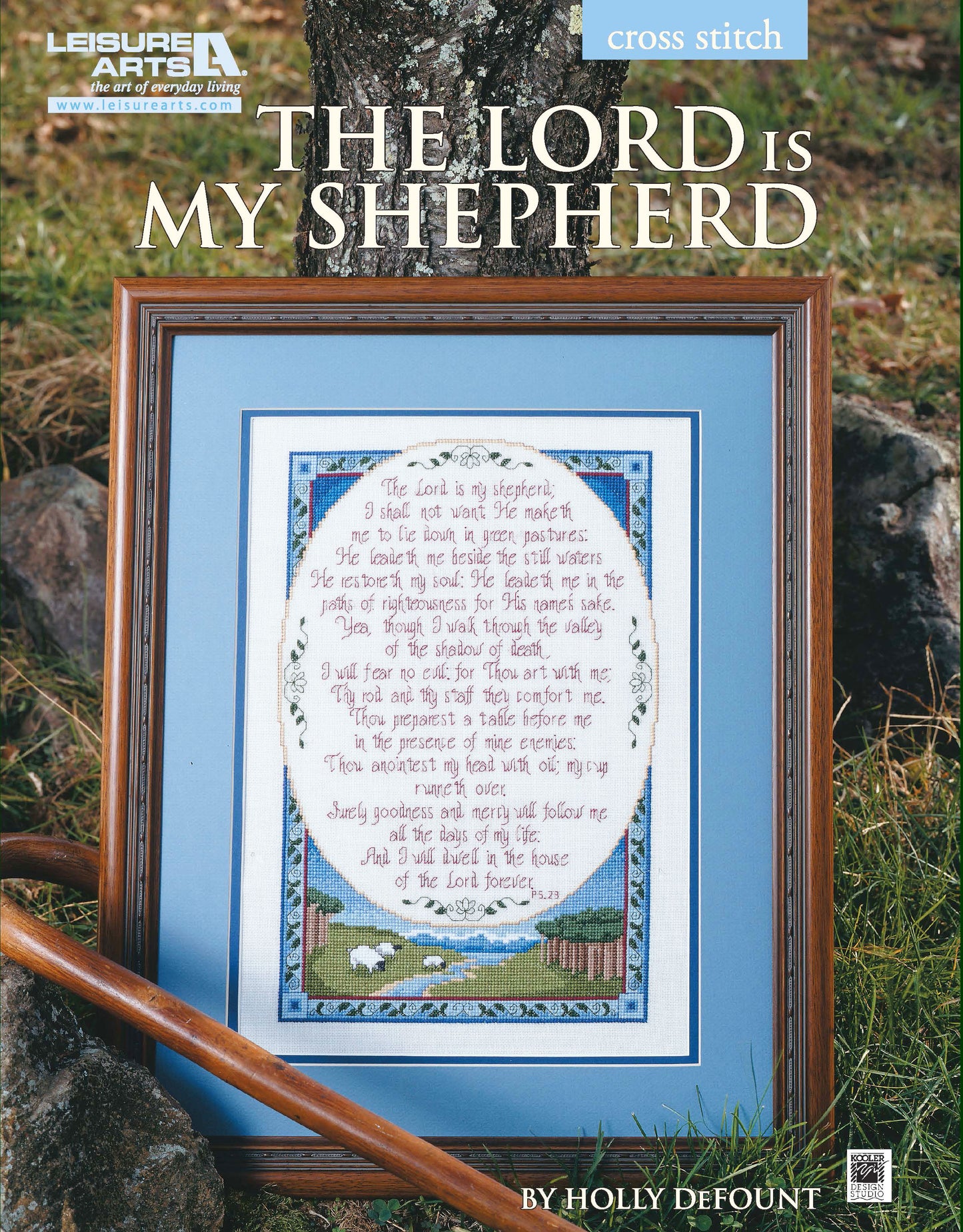 The Leisure Arts "The Lord is My Shepherd" framed cross stitch features a blue border, pastoral scene with sheep, and the Bible verse in large text, displayed against a tree and grass.