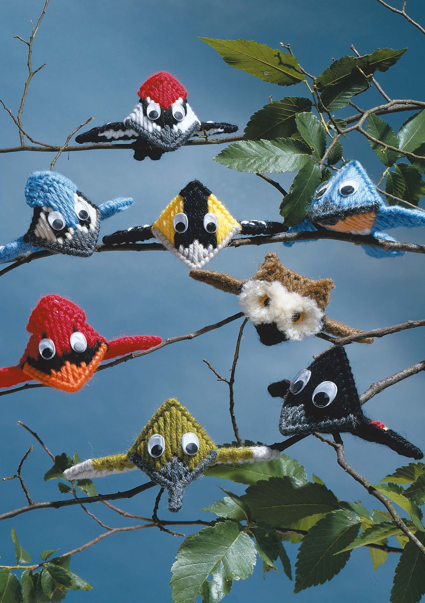 Several colorful Big Beak Birds in Plastic Canvas by Leisure Arts perch on tree branches with big eyes, set against a blue sky and green leaves. These cute designs are ideal for beginners looking to brighten up any space.