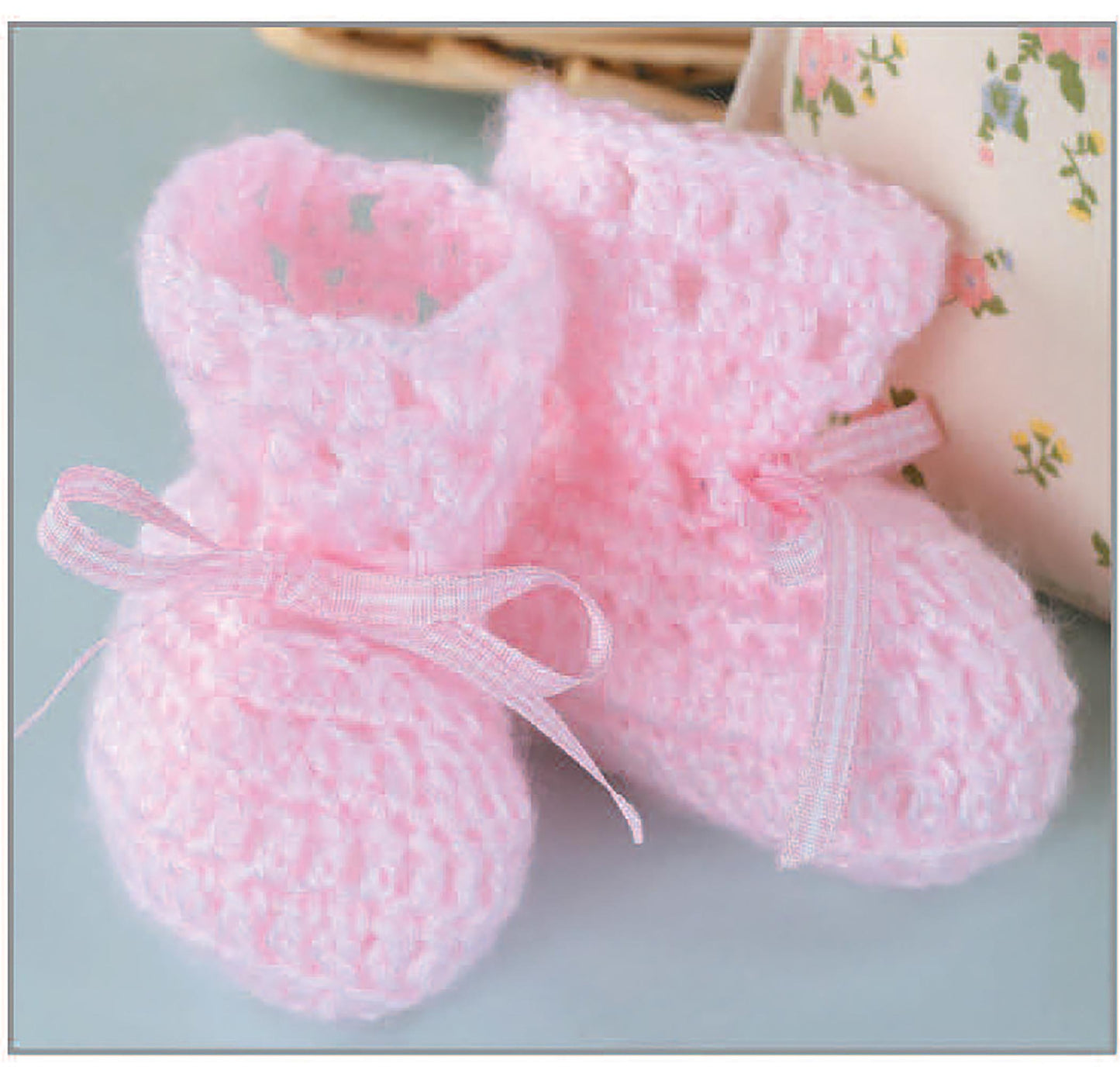A pair of soft, pink, knitted baby booties with ribbon bows sit side by side on a light surface. In the background is a floral-patterned afghan, made from patterns in "Baby Afghans" by Leisure Arts.