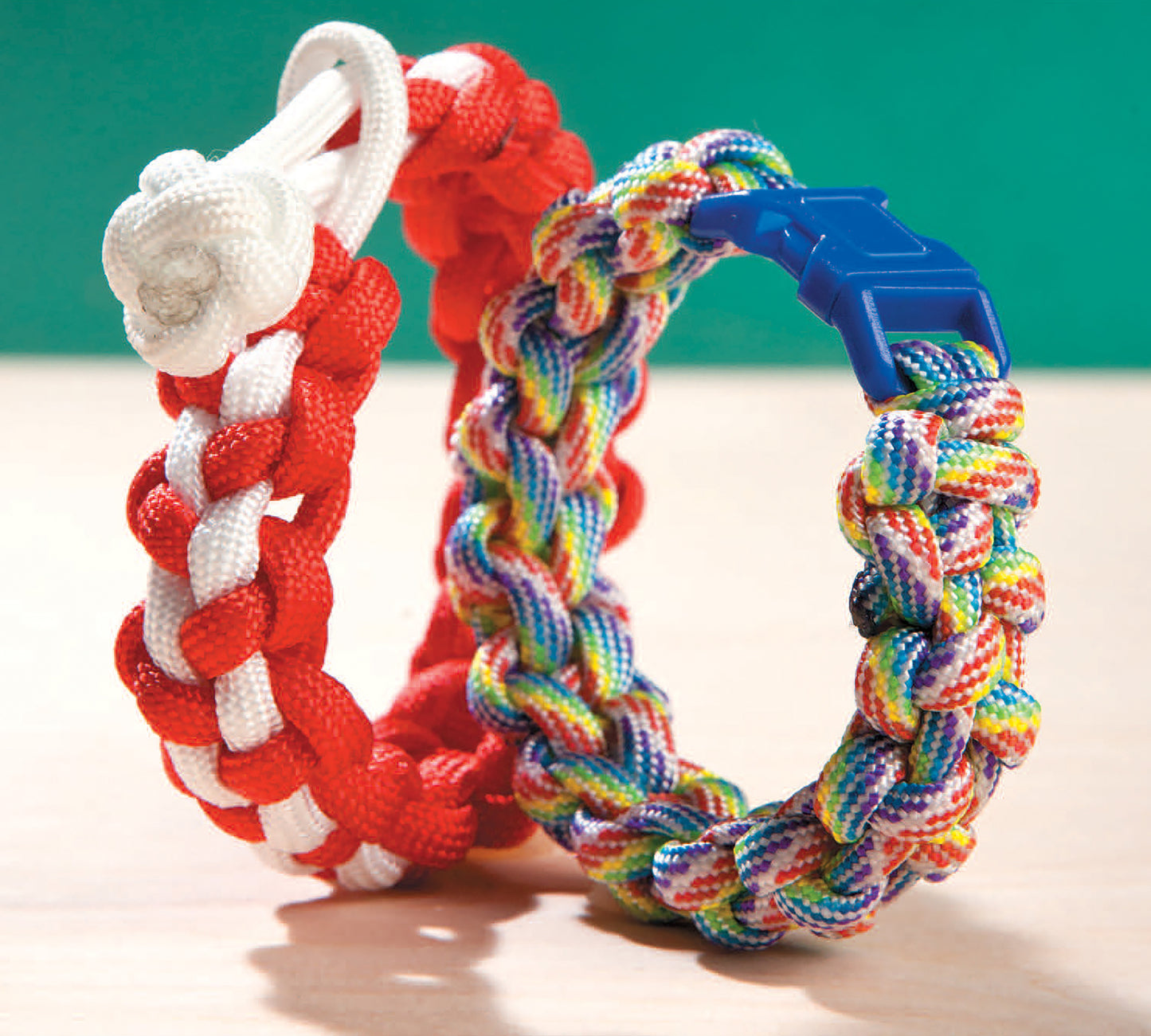 Two stylish Paracord Crafts bracelets from Leisure Arts—one red and white with a knot closure, the other multicolored with a blue clasp—feature classic cobra stitch and are displayed upright against a green background.