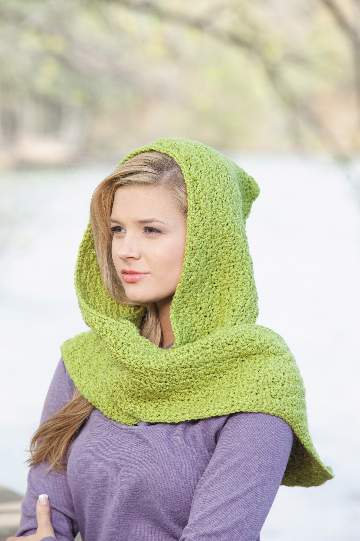 A woman stands outdoors by a river wearing a textured, bright green scarf from "Hooded Scarves, Book 2 Digital Download" by Leisure Arts over a purple sweater, with trees and water blurred in the background.