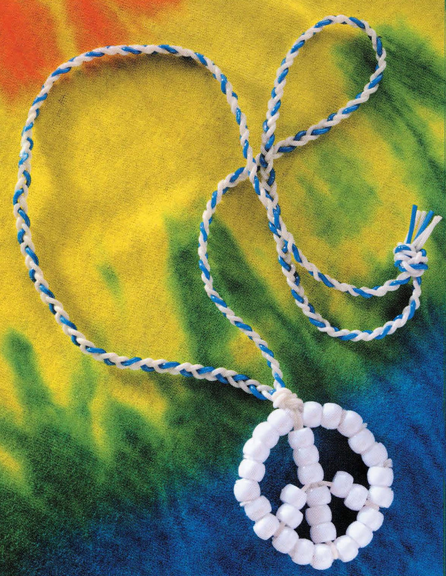 A Plastic Lacing & Pony Beads necklace by Leisure Arts, featuring blue and white braids and a beaded peace sign pendant, rests on vibrant tie-dye fabric, highlighting its unique lacing style.