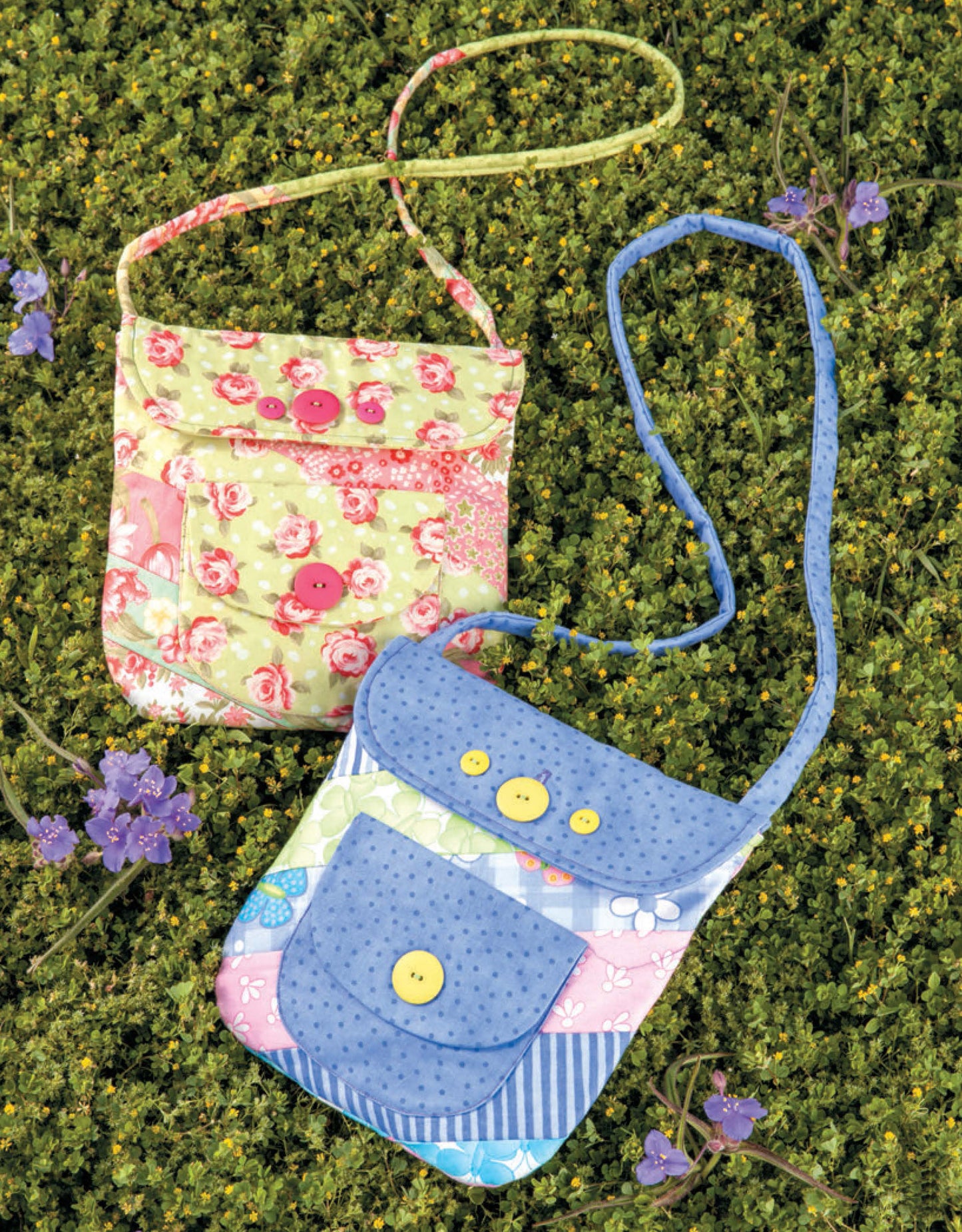 Two small purses made from fat quarters, featured in Sew Totes & Bags Digital Download by Leisure Arts, rest on grass with purple flowers—one is pink and floral, the other blue with patchwork patterns and both have button closures.
