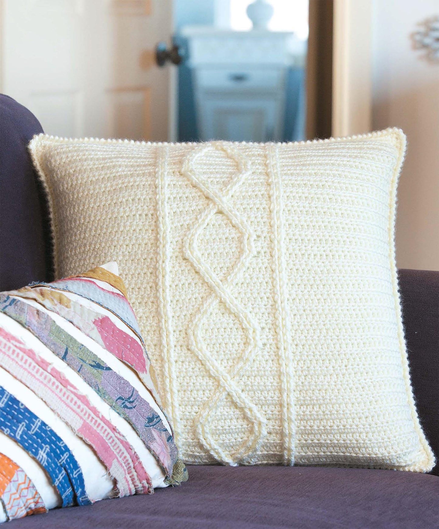 A white pillow with classic crochet cable patterns, inspired by "Beginner's Guide to Crochet Cables" from Leisure Arts, rests on a dark couch beside a vibrant striped pillow. A doorway and white cabinet appear in the background.