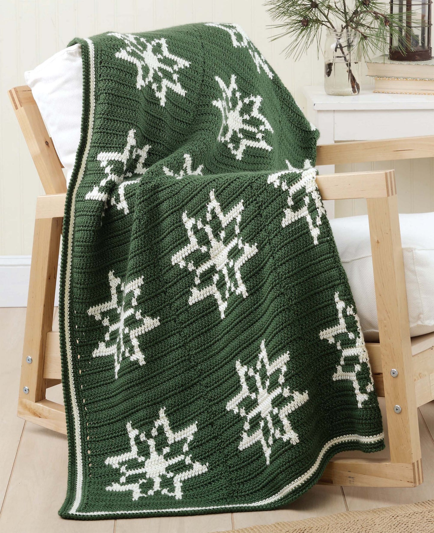 A green blanket with Fair Isle crochet snowflakes, draped on a chair, is made using patterns from Leisure Arts’ "Beginner's Guide to Crochet Color Work." A vase with pine branches decorates a nearby table.