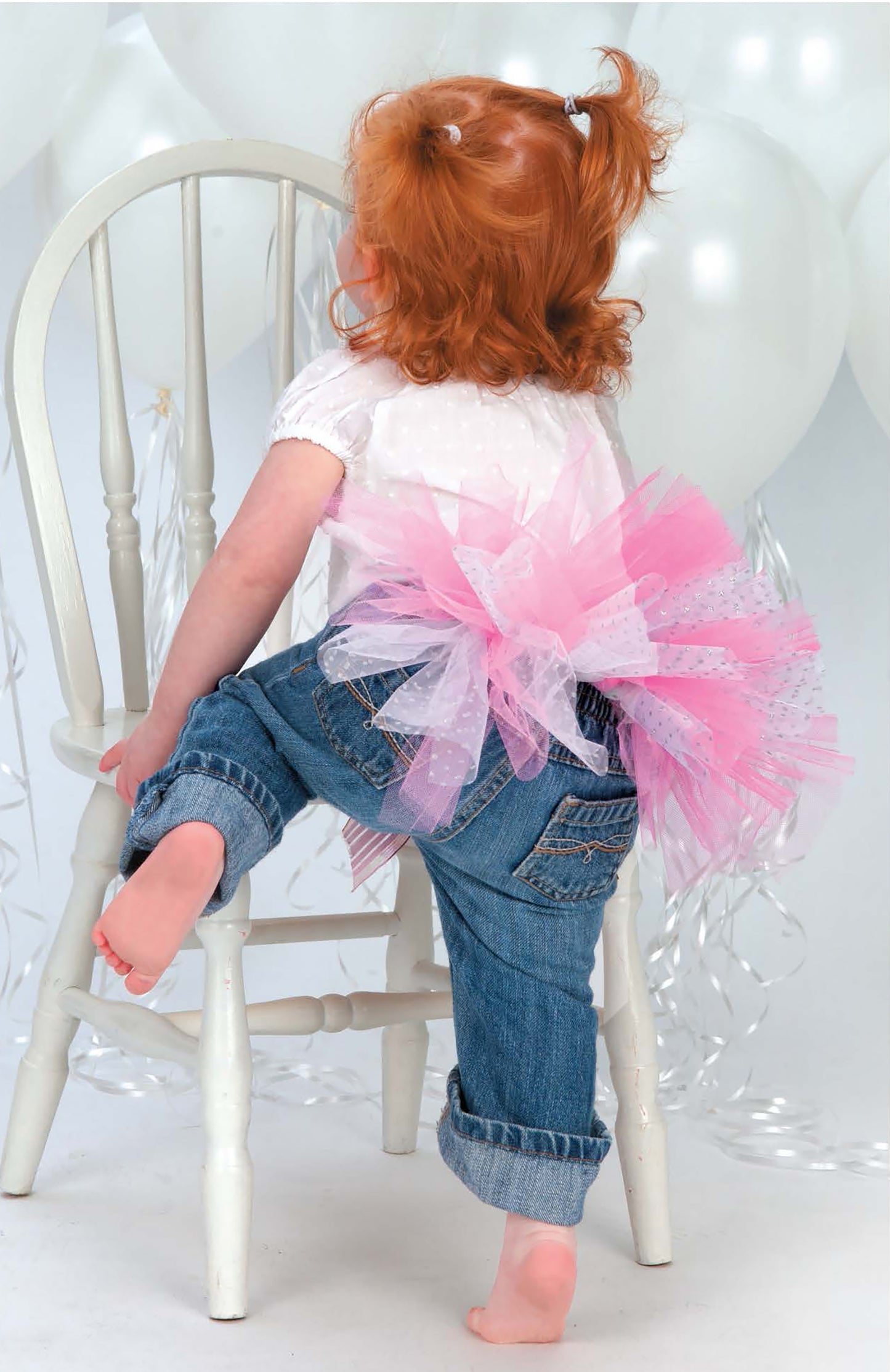 A red-haired young child in blue jeans, a white shirt, and Leisure Arts’ "A Tutu for Every Occasion" pink tutu climbs barefoot onto a white chair. White balloons float in the background, creating a playful, festive scene.