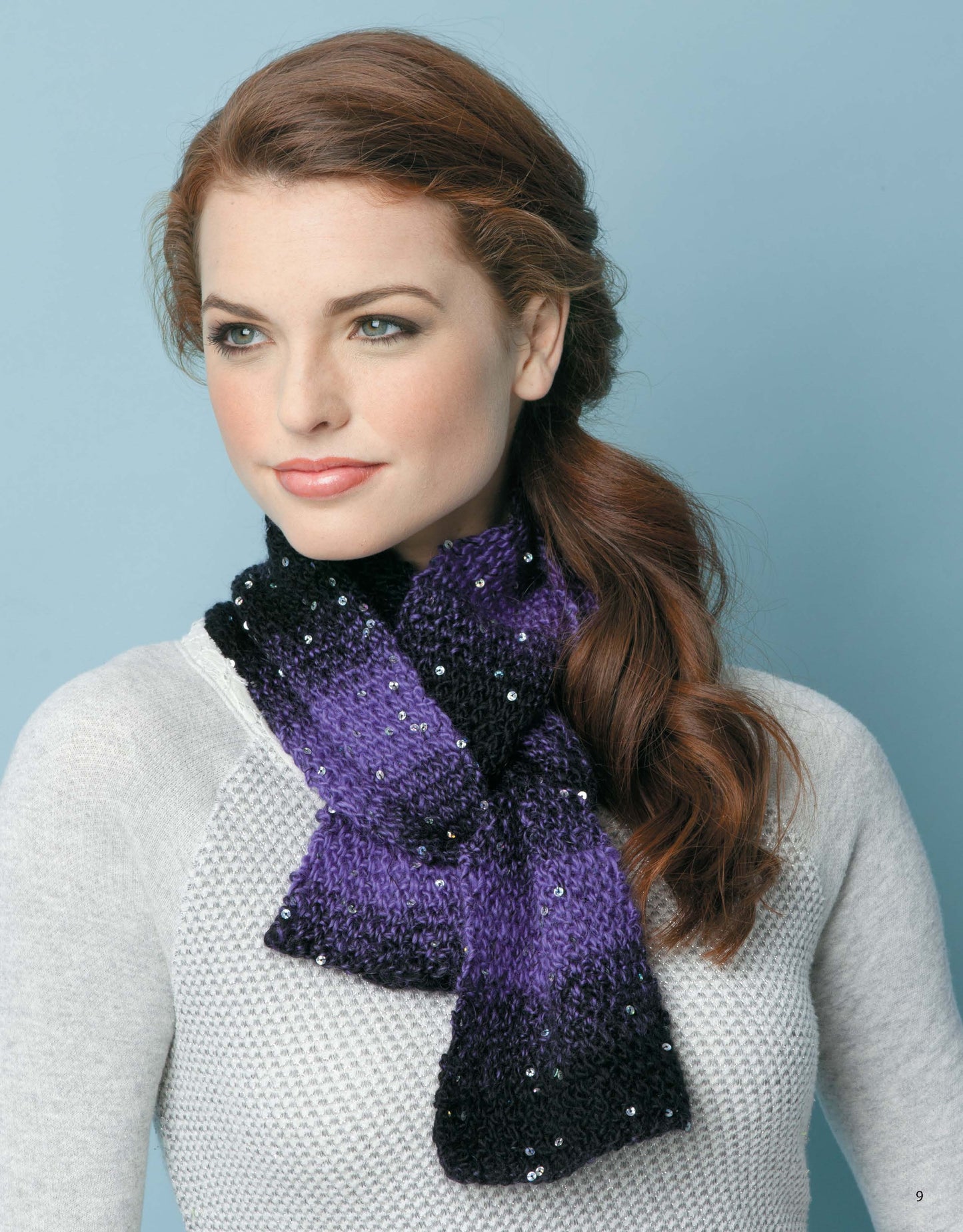A woman with wavy auburn hair models a purple and black sequin scarf from Leisure Arts’ Knit Scarves & Cowls collection, inspiring beginners or anyone seeking new scarf patterns, set against a light blue background.